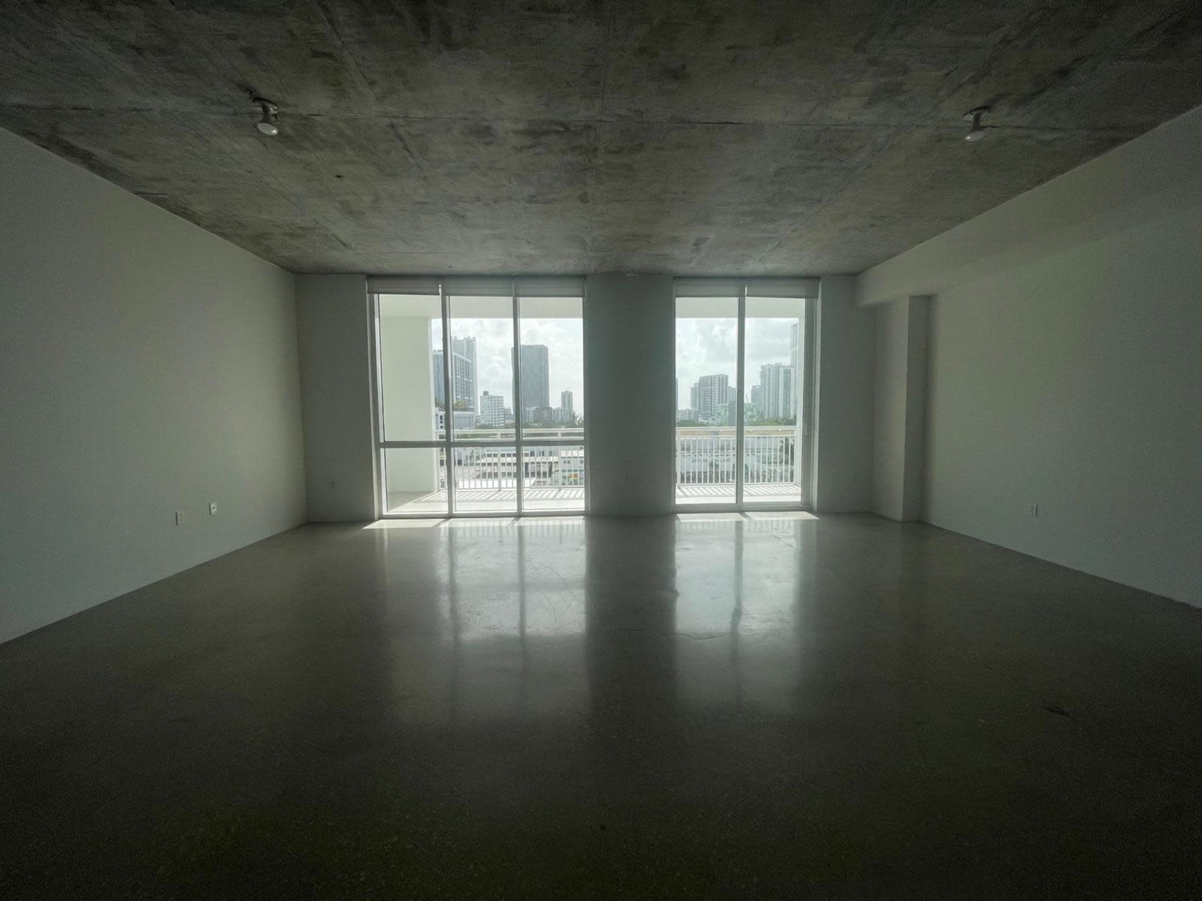 The image shows a spacious, empty room with large windows that offer a view of a city skyline.
