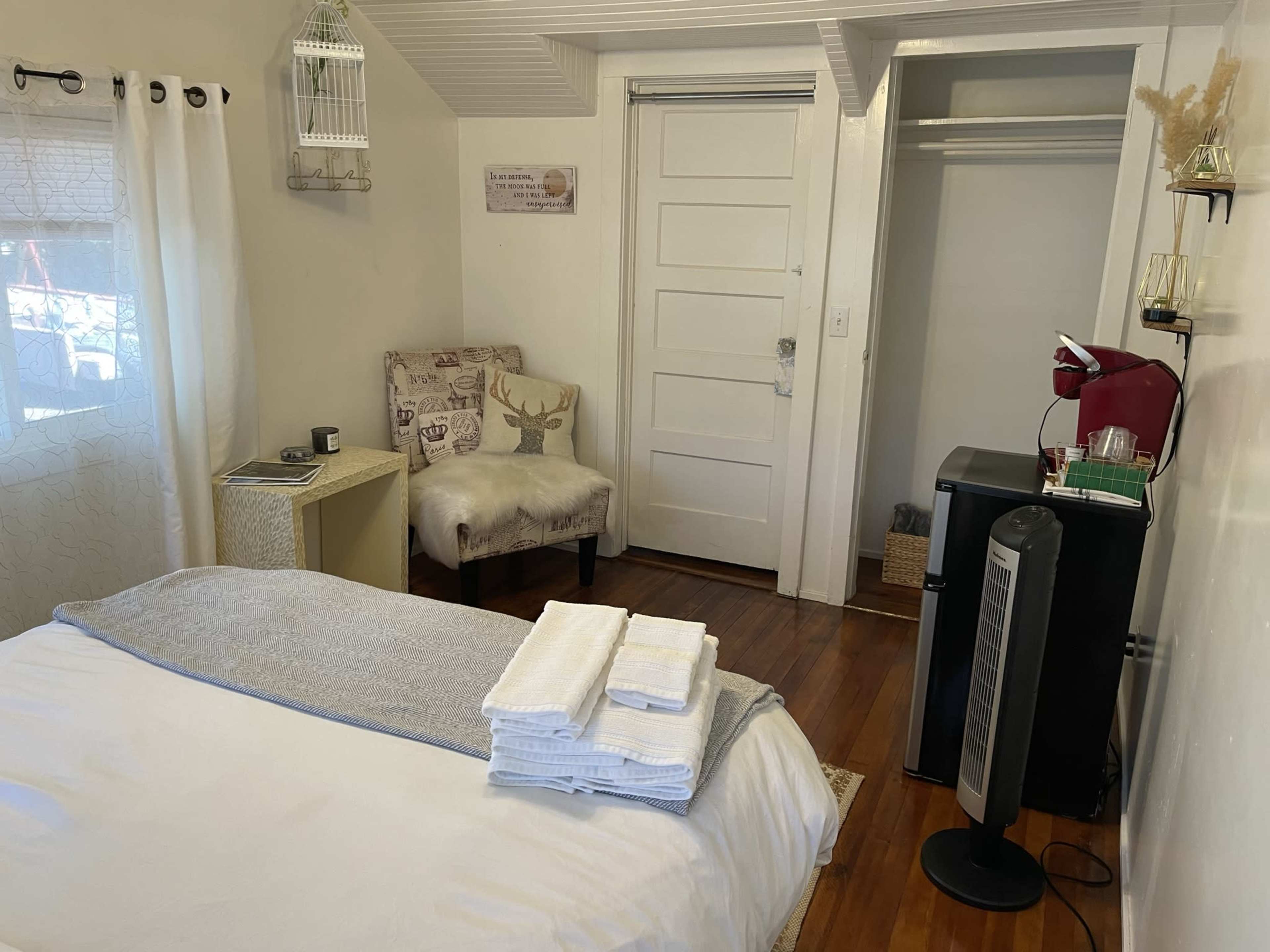 A small, well-organized room featuring a bed with neatly folded towels, a chair with a deer design, a small table, a closet, and a coffee machine.