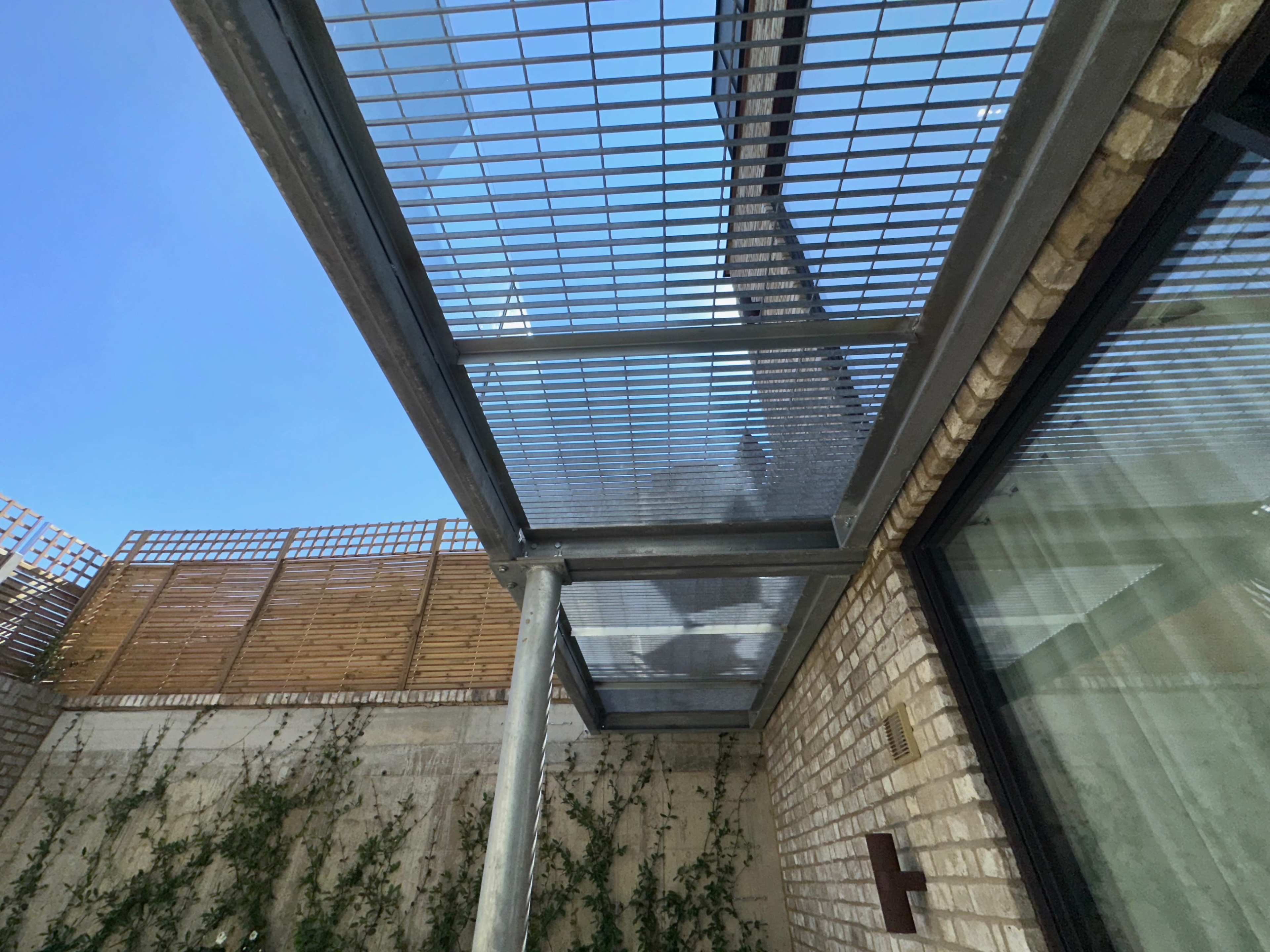 The image shows a steel grid canopy covering a small outdoor space, with a wall of brick and greenery visible below.
