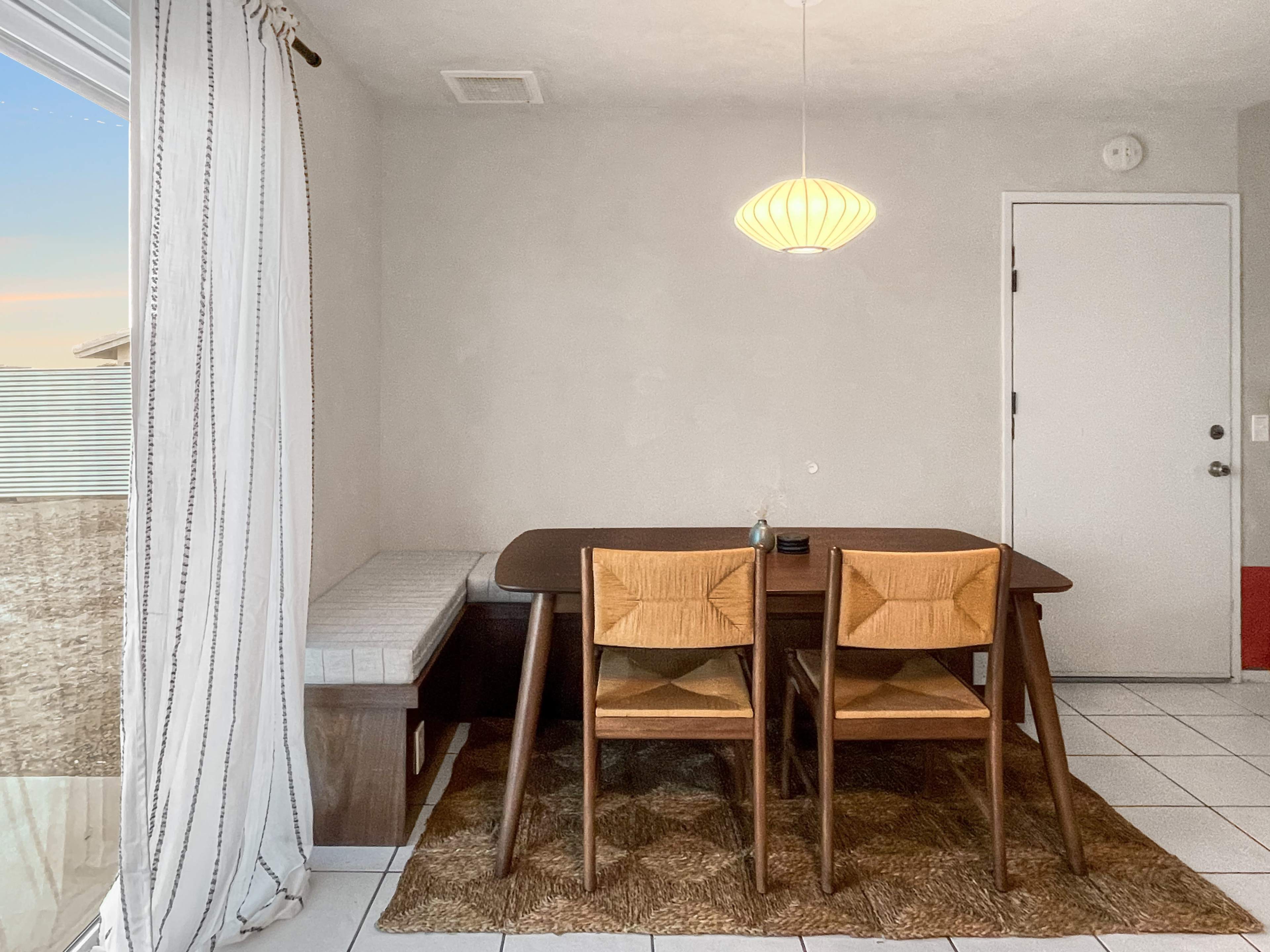 A wooden dining table with two chairs is set in a brightly lit room, next to a window with sheer curtains and a door leading outside.