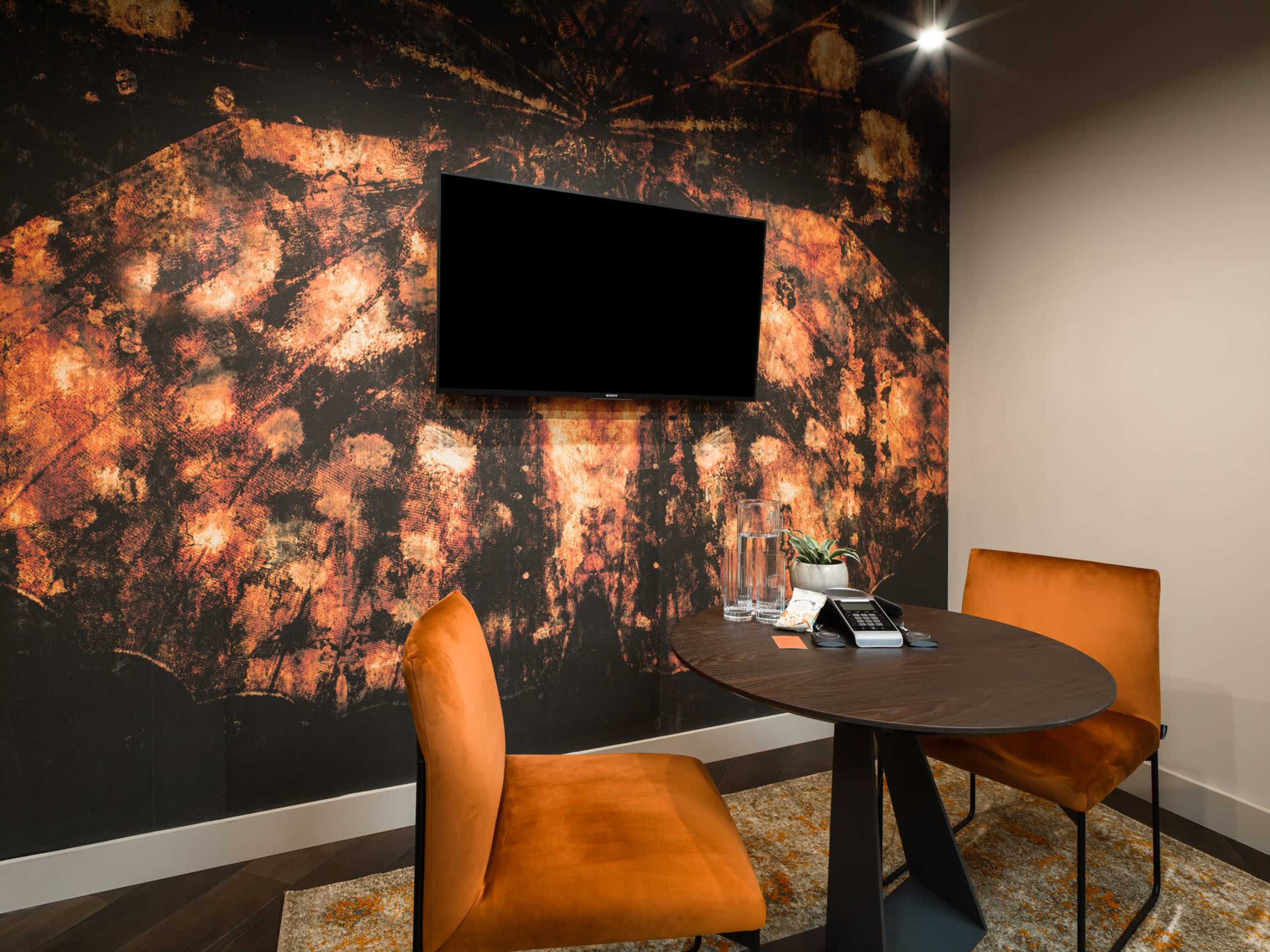 A small meeting room features a dark wall with an abstract patterned wallpaper, a round table with two orange chairs, and a wall-mounted television.