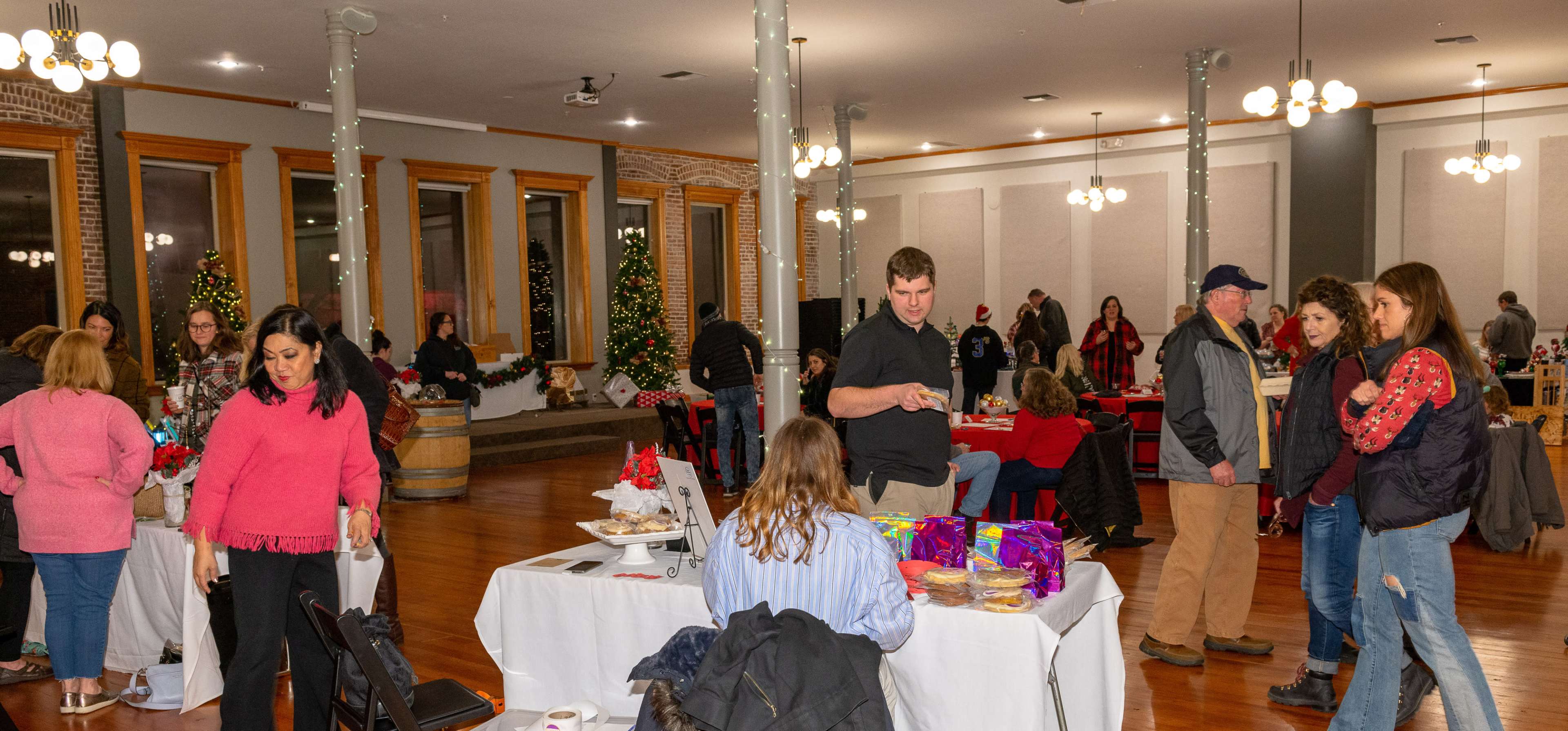 A community event is taking place in a spacious hall decorated with Christmas trees, where attendees are browsing tables with festive displays and treats.