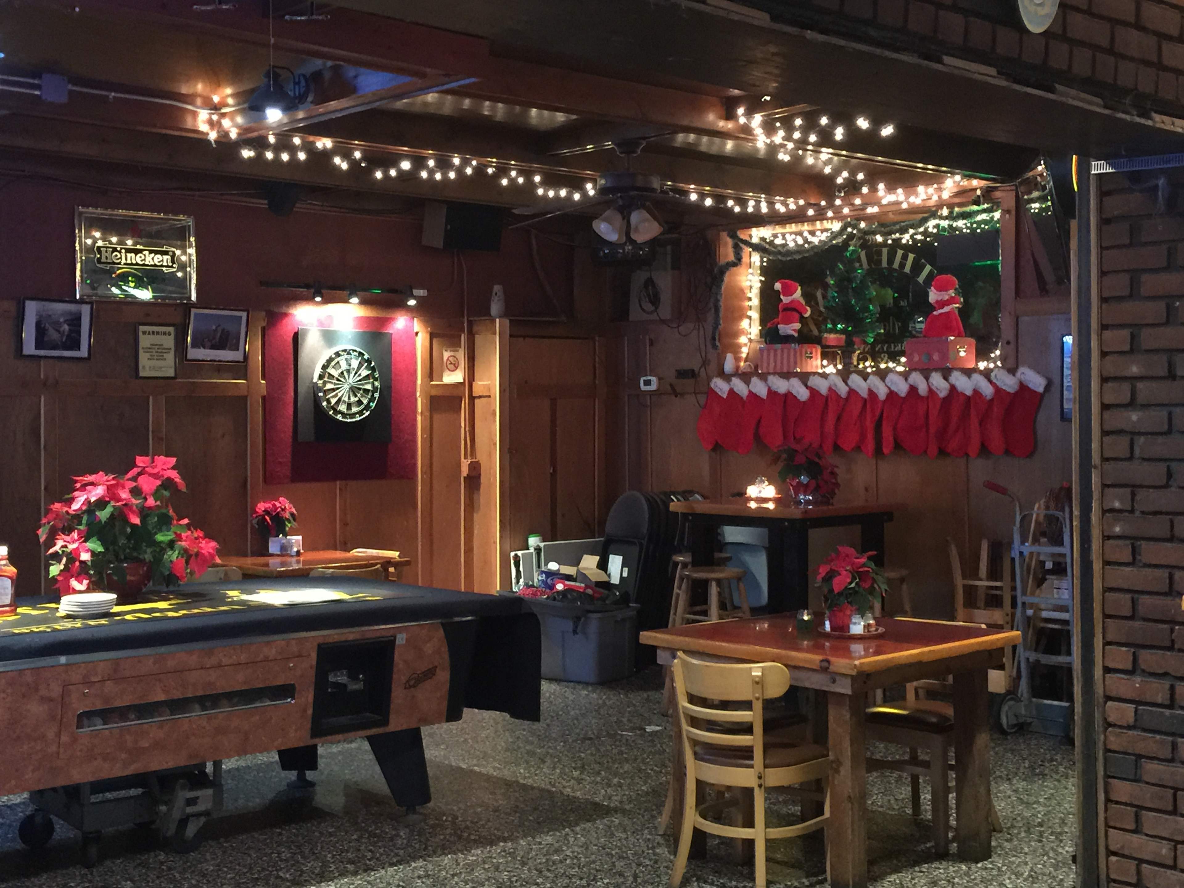 The interior of a bar features a dartboard area, decorated with string lights and stockings, along with tables set with poinsettias.