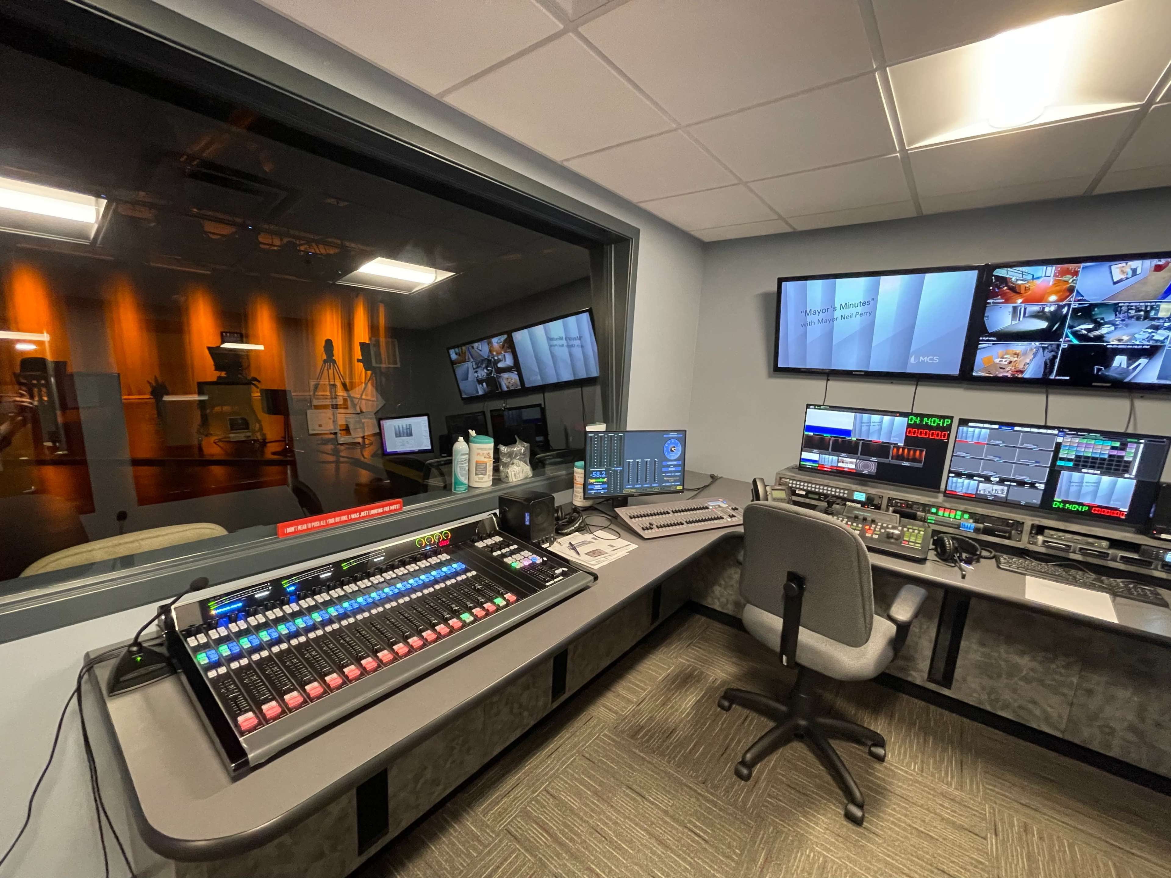 A television control room with a main mixing console, multiple screens displaying various feeds, and a view into a production area.
