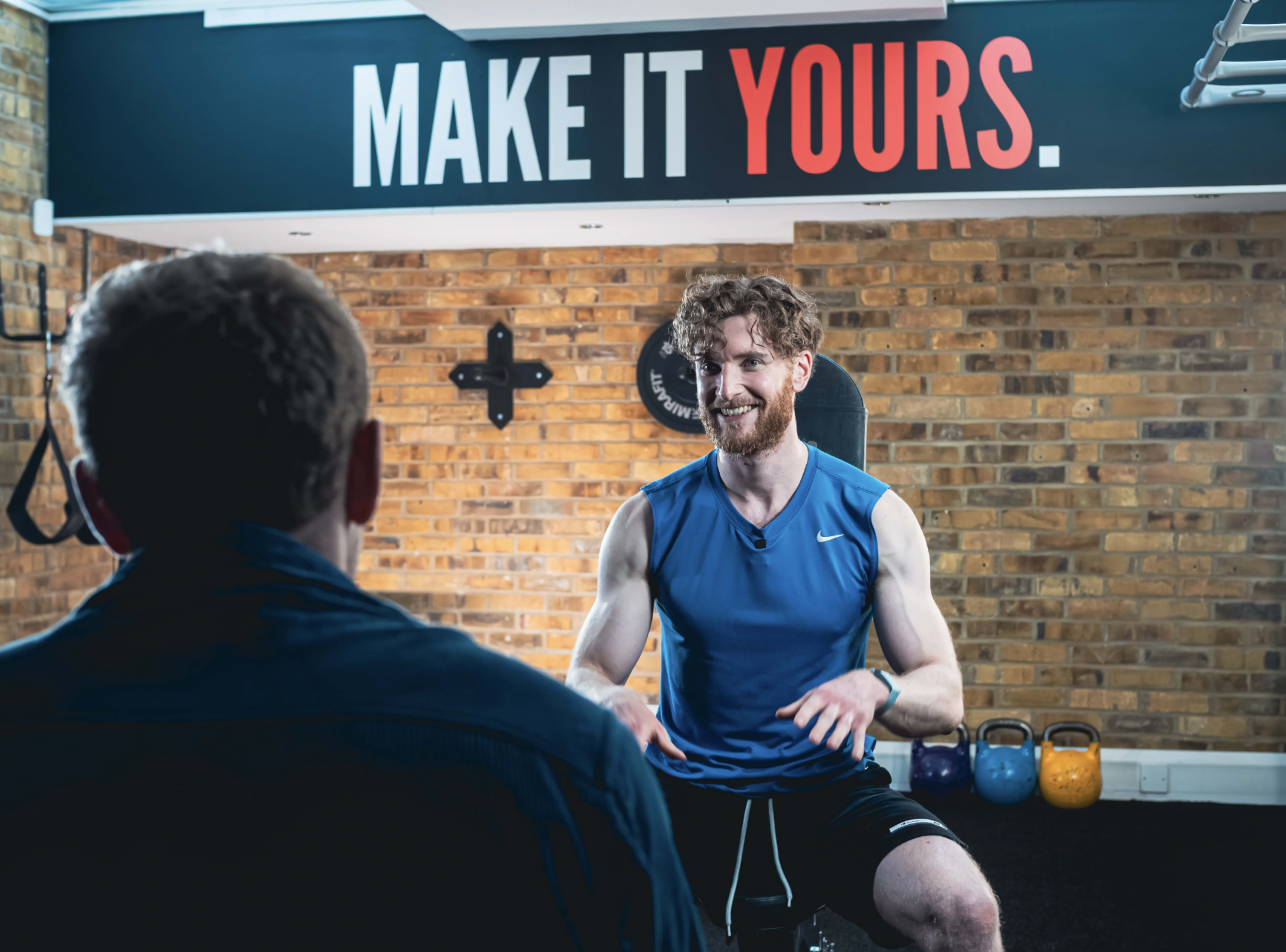 A man in a blue sleeveless shirt smiles while seated in a gym, facing another person out of view, against a wall with the text "MAKE IT YOURS."
