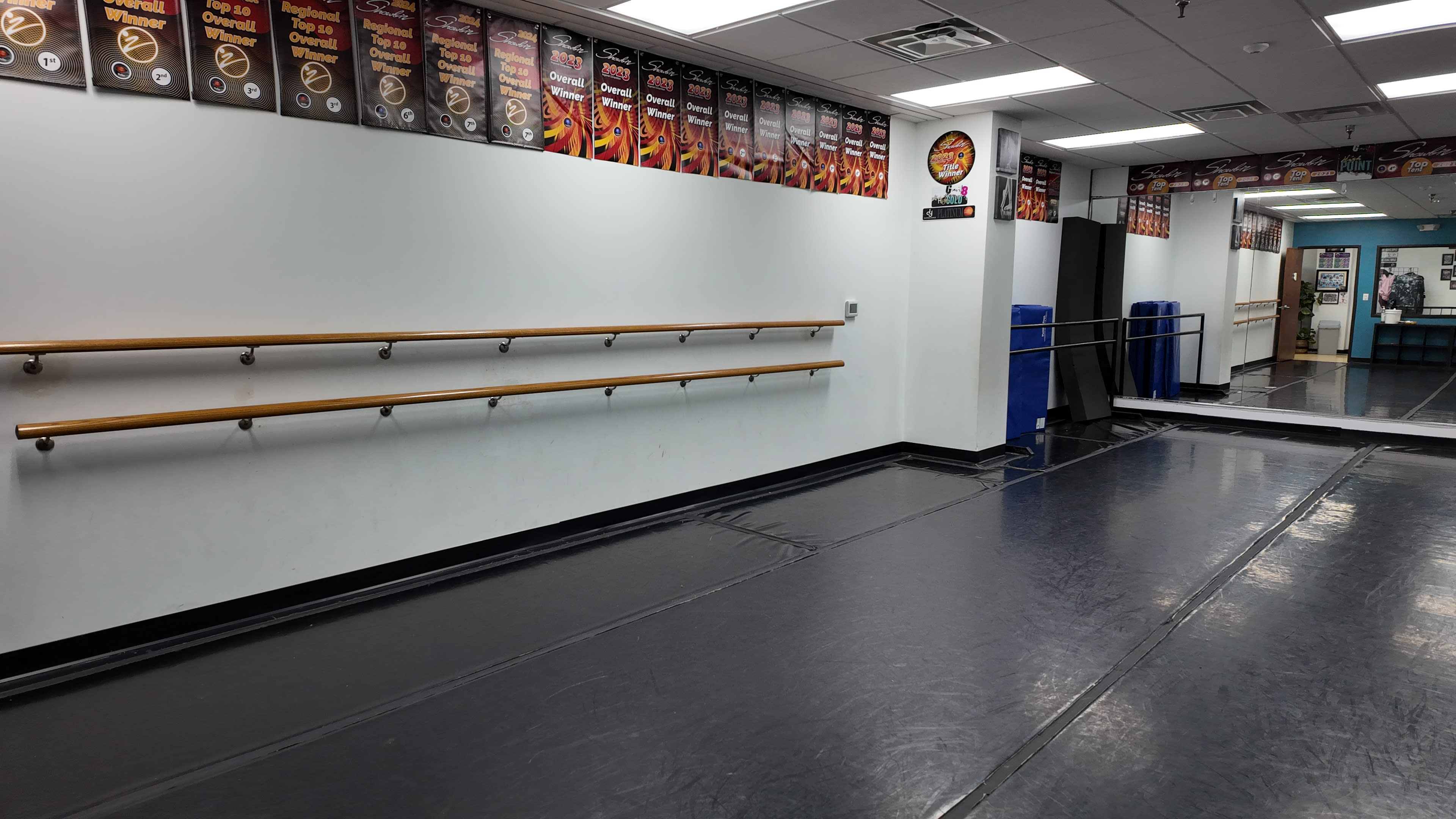 The image shows a dance studio with wooden ballet bars along one wall and mirrors on another, featuring a black floor and several posters hanging above the bars.