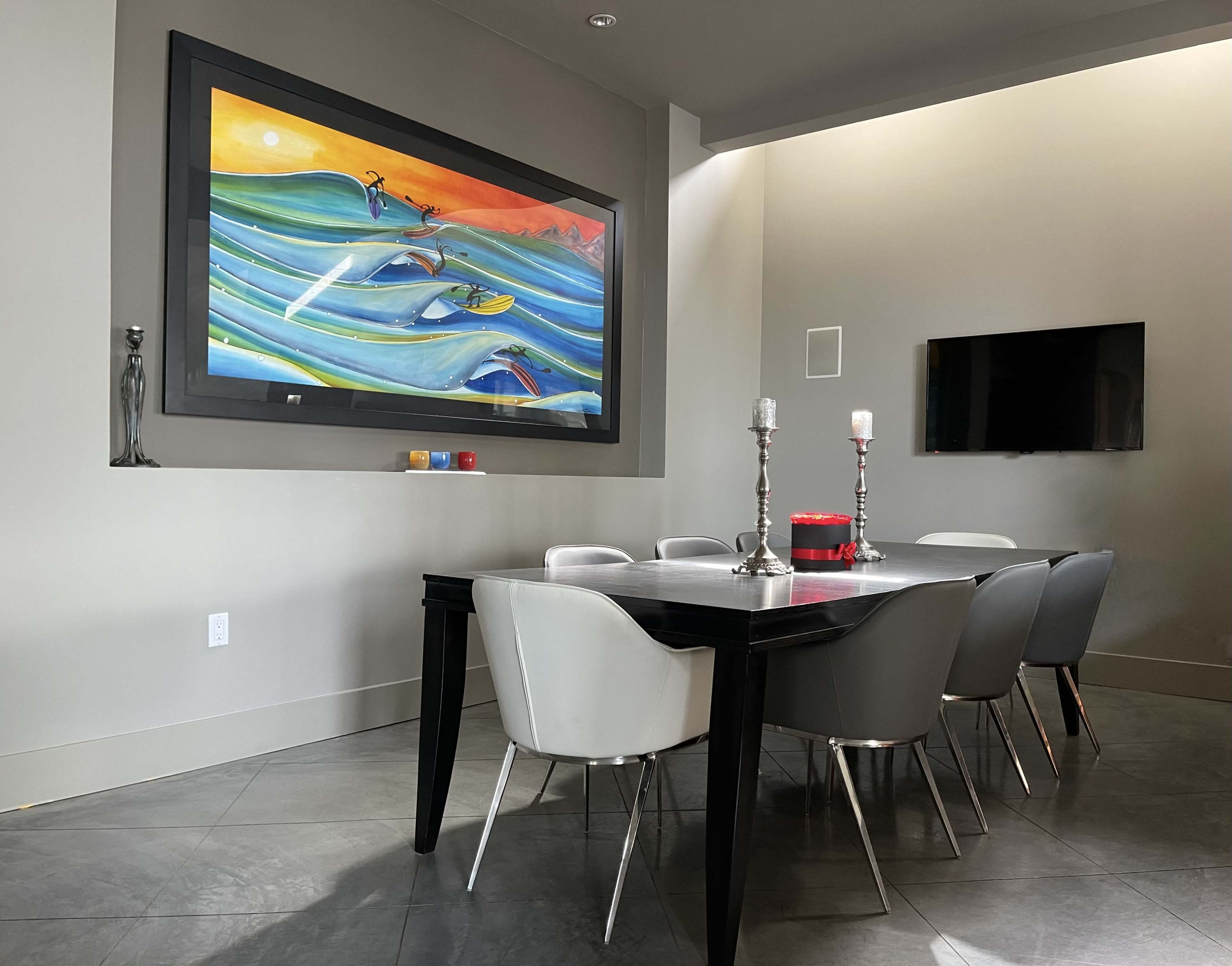 A modern dining area featuring a large table surrounded by chairs, with a colorful ocean-themed painting on the wall and a TV mounted nearby.