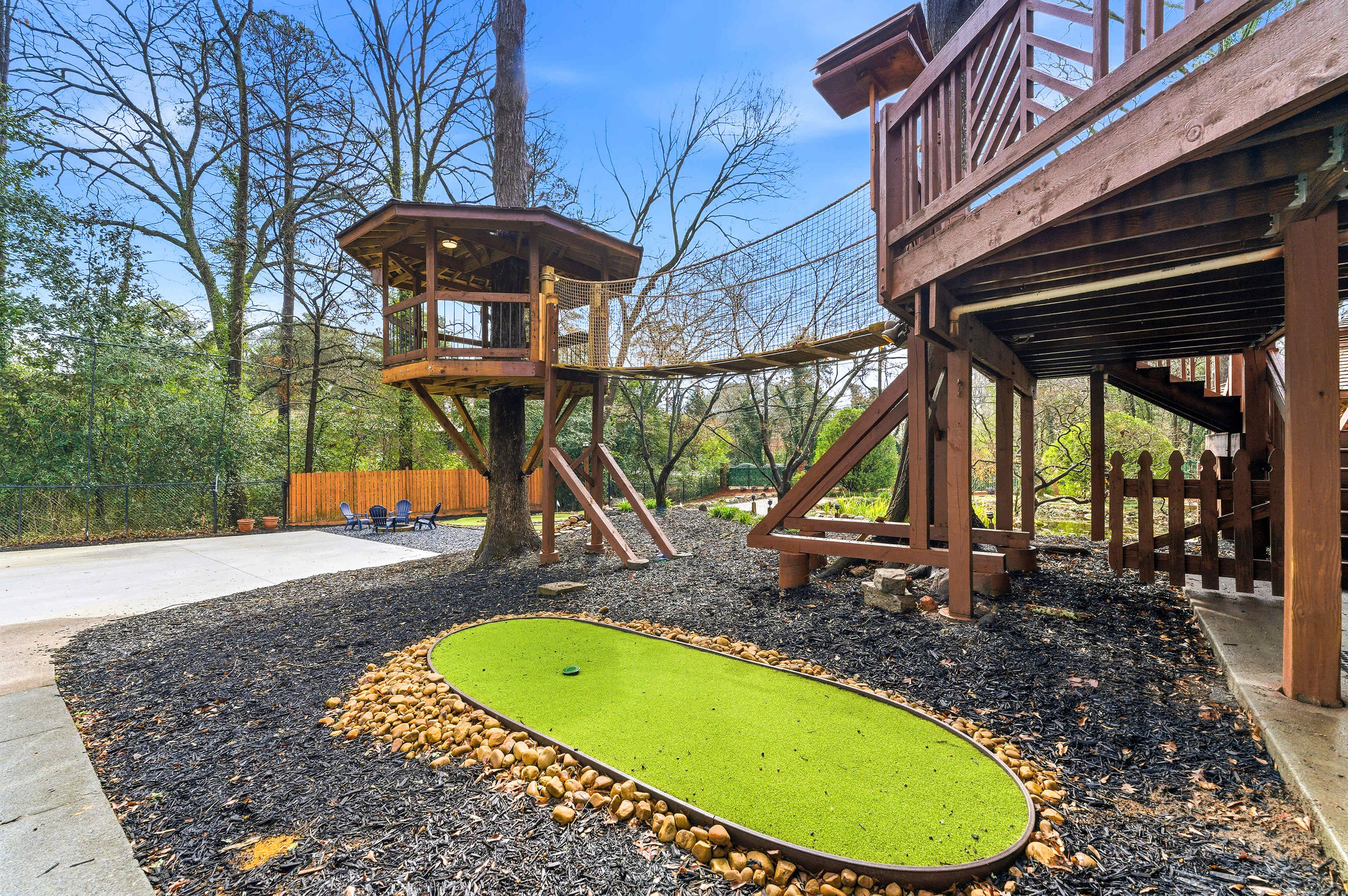 The image shows a wooden play structure with a climbing area and a netted walkway, alongside a small artificial putting green surrounded by pebbles and mulch.