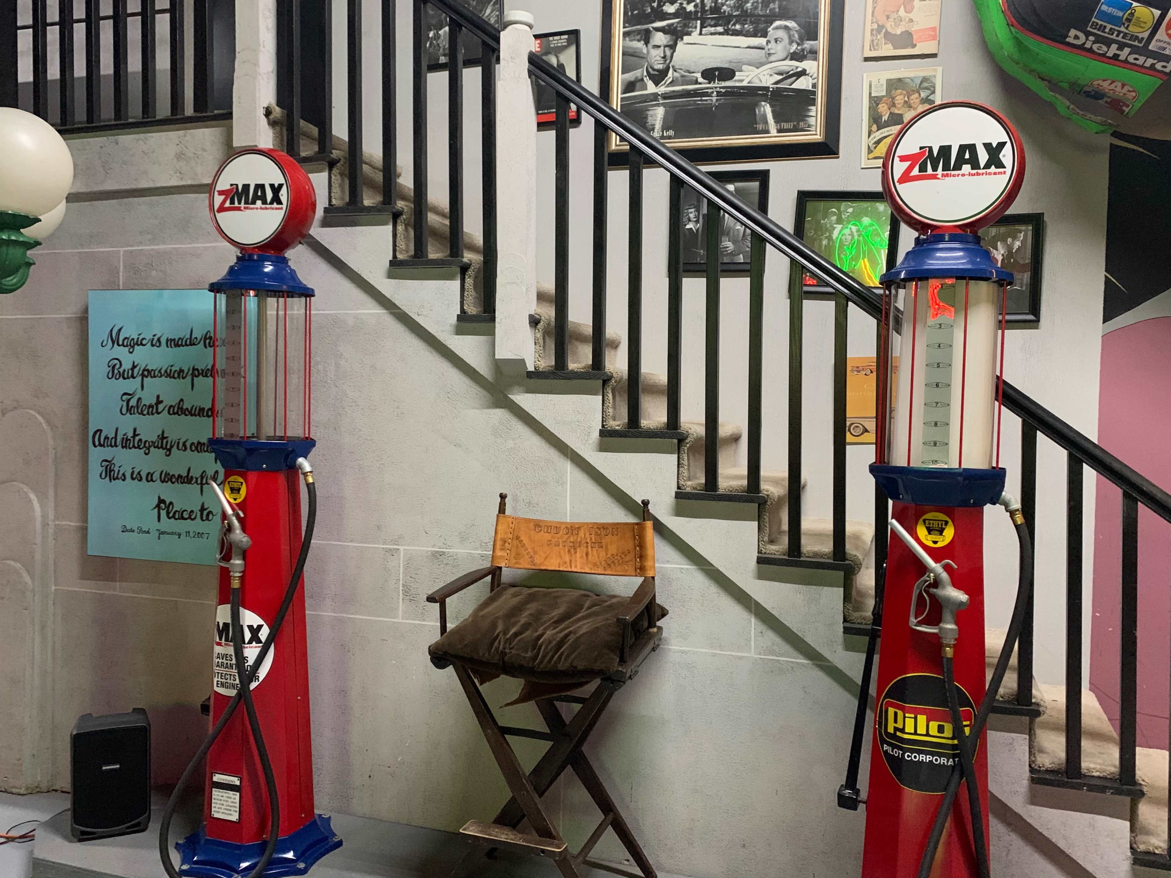 Two vintage gas pumps flanking a wooden director's chair at the bottom of a stairway, with framed photographs on the wall above.