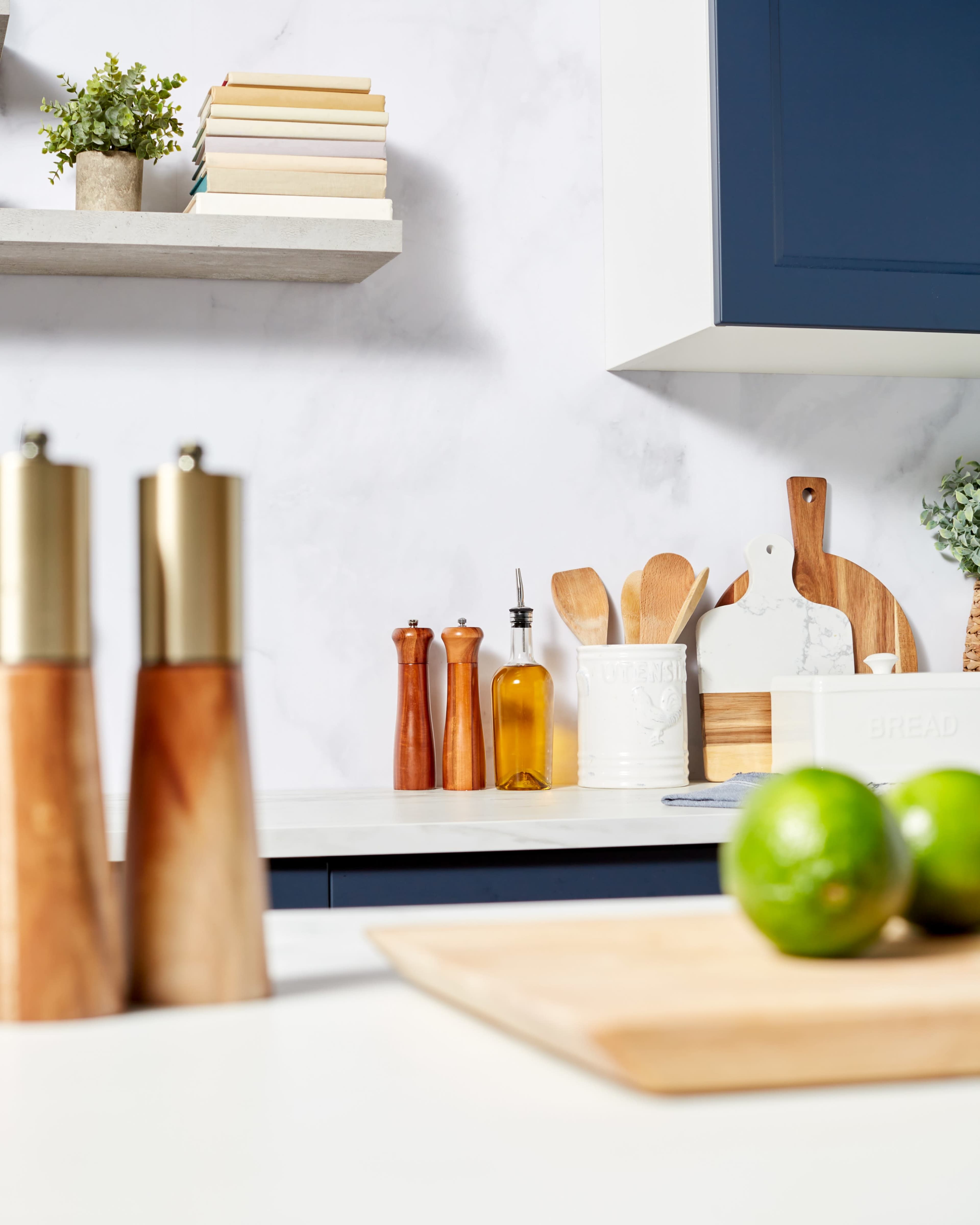 A modern kitchen countertop with wooden utensils, spices, an olive oil bottle, and fresh limes on the surface, accompanied by a shelf displaying stacked books and a potted plant.