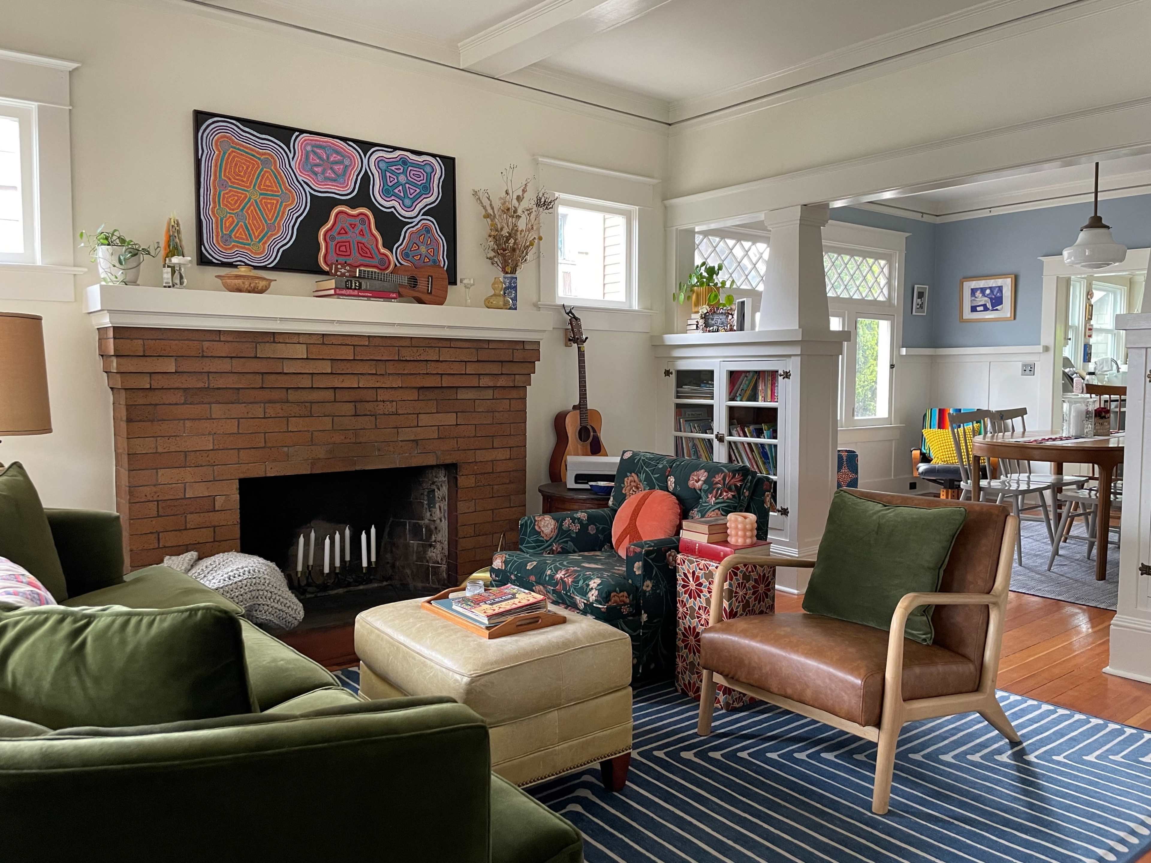 A living room with a brick fireplace, green couches, a floral armchair, a guitar, and a dining area in the background.