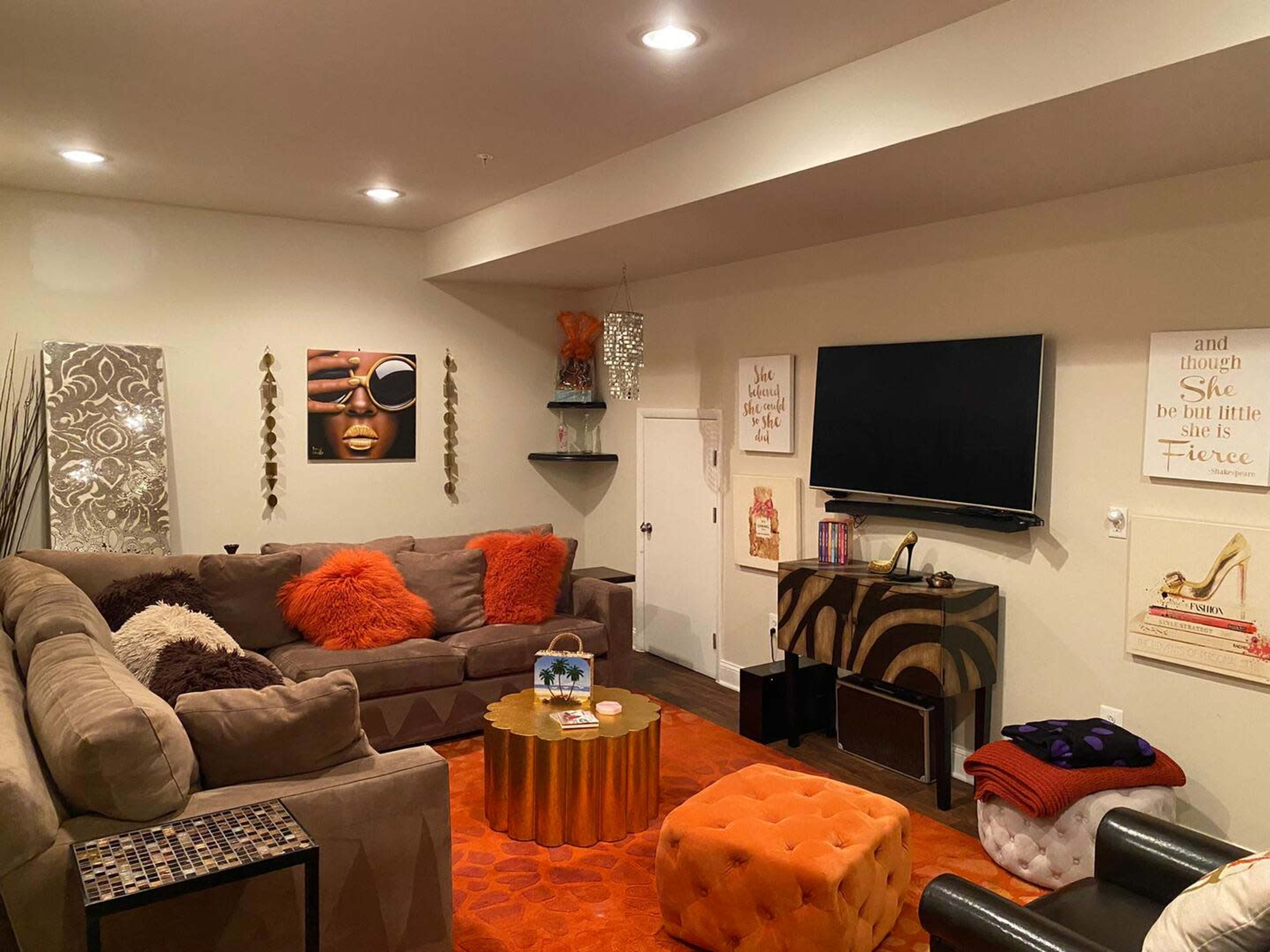 The image shows a cozy living room with a brown sectional sofa, a round coffee table, colorful ottomans, a television mounted on the wall, and decorative artwork.