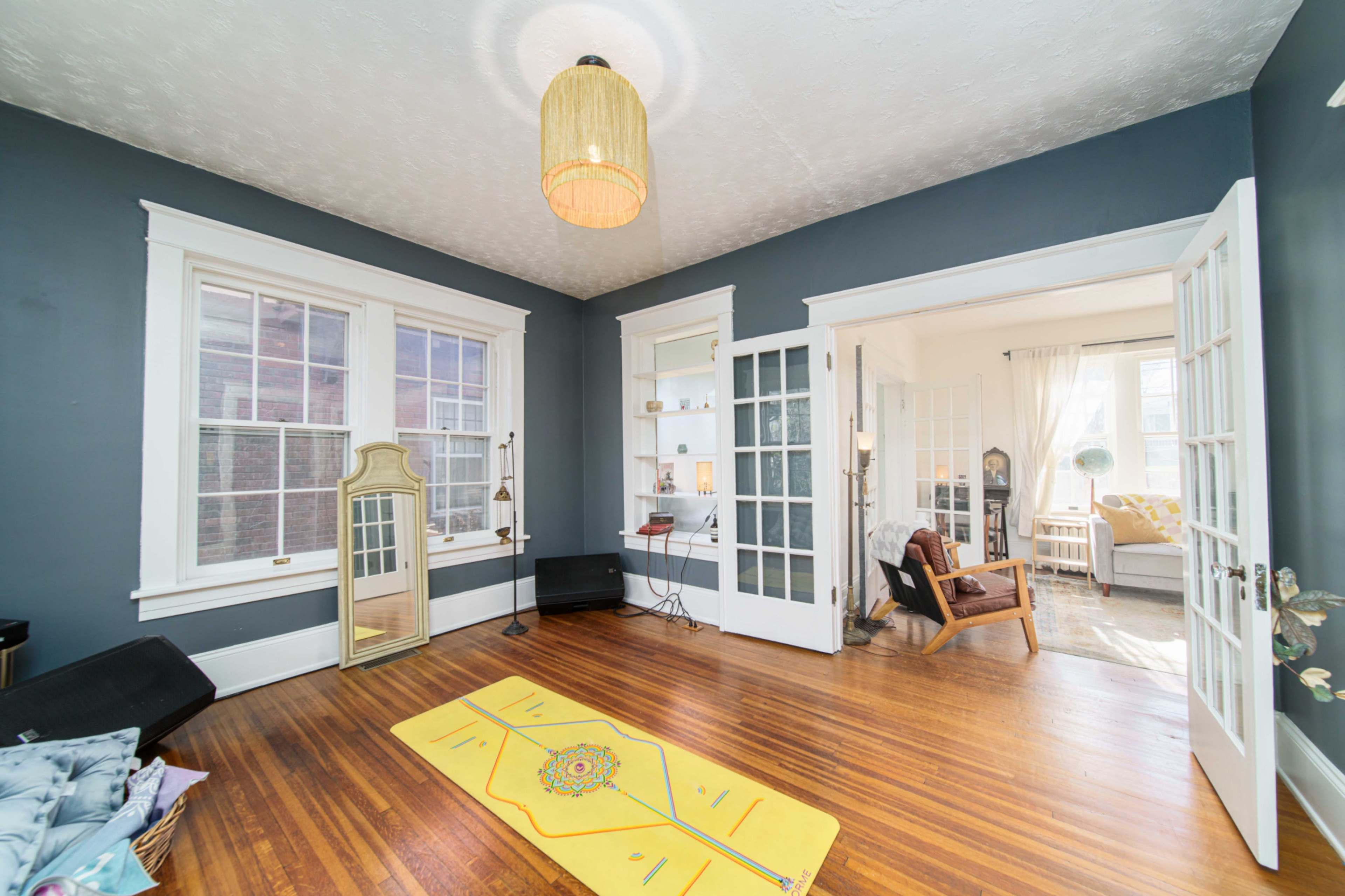 The image depicts a bright living room with hardwood floors, large windows, and a cozy seating area separated by glass doors.