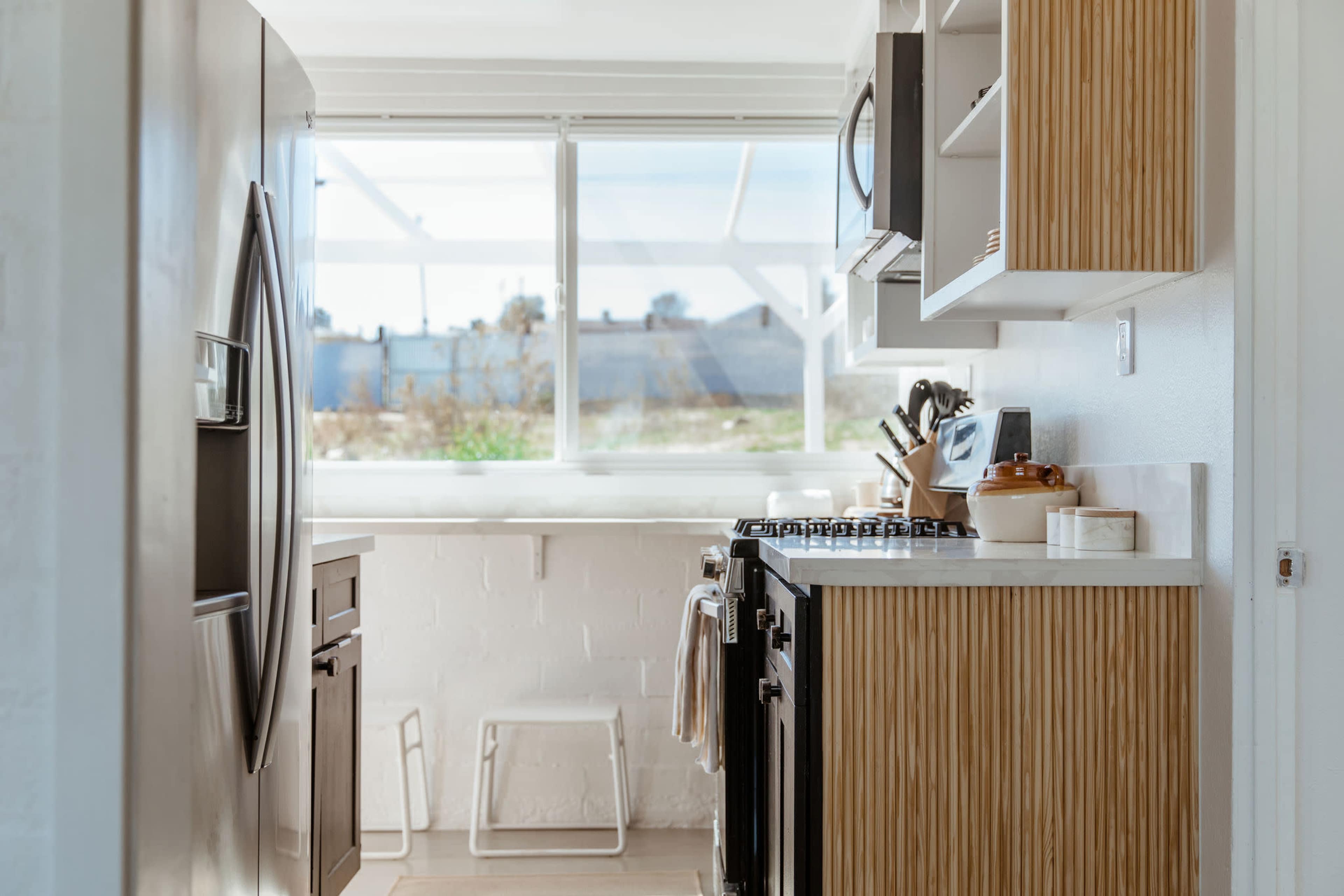 A modern kitchen features stainless steel appliances, a white countertop, and wooden cabinetry, with windows revealing an outdoor view.