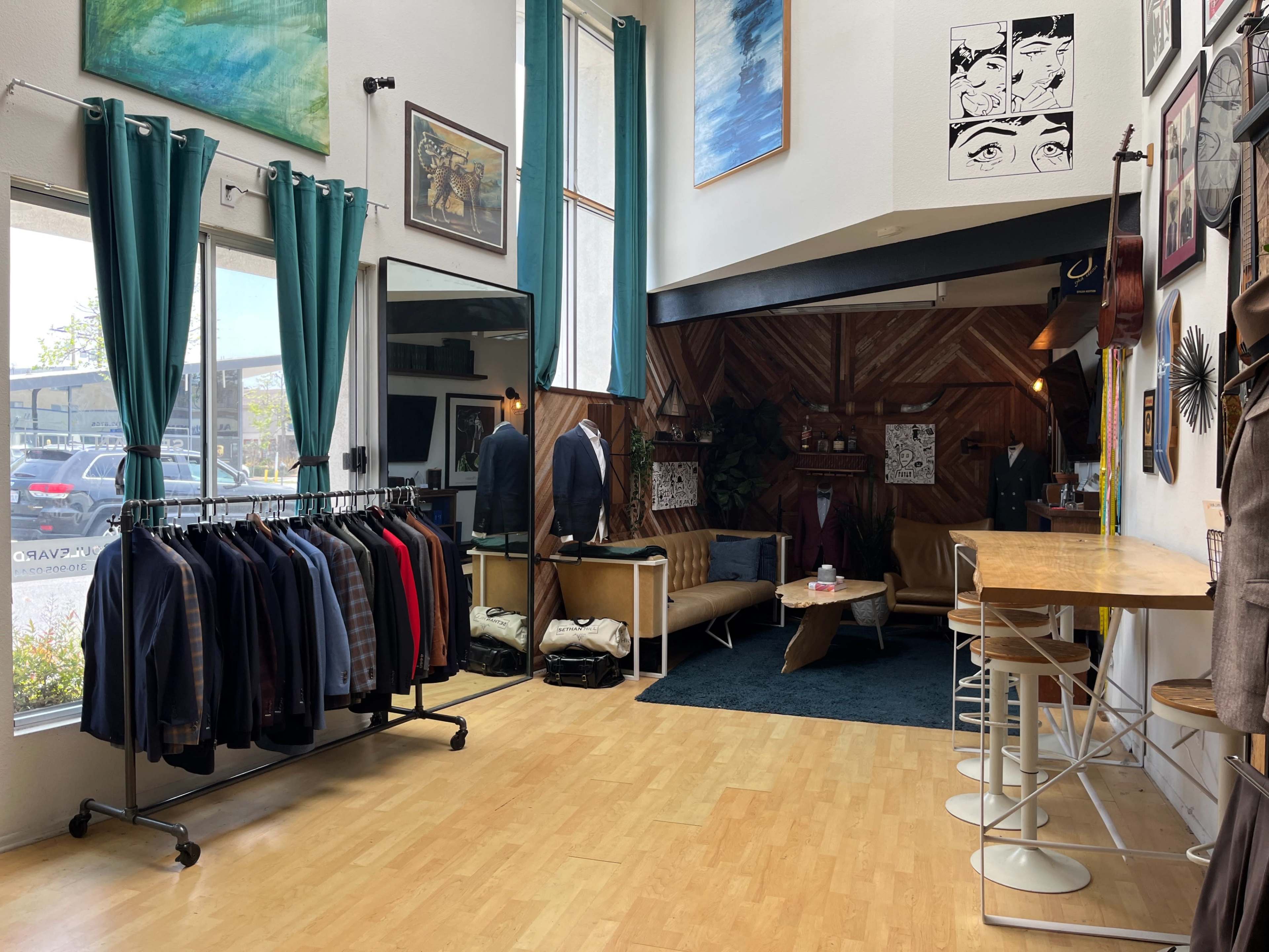 A boutique interior featuring a clothing rack with suits, a lounge area with a couch and chairs, and various artworks on the walls.