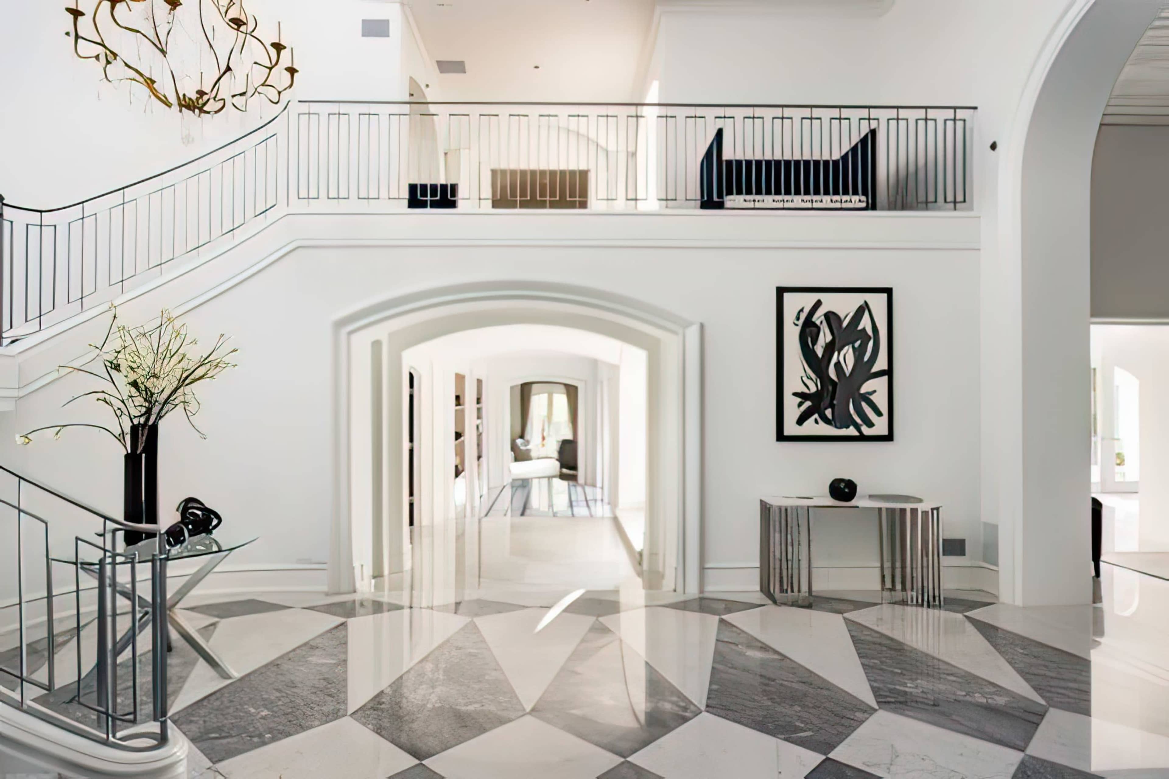 A spacious, modern interior with a white staircase, a geometric marble floor, and an arched entryway leading to a hallway.