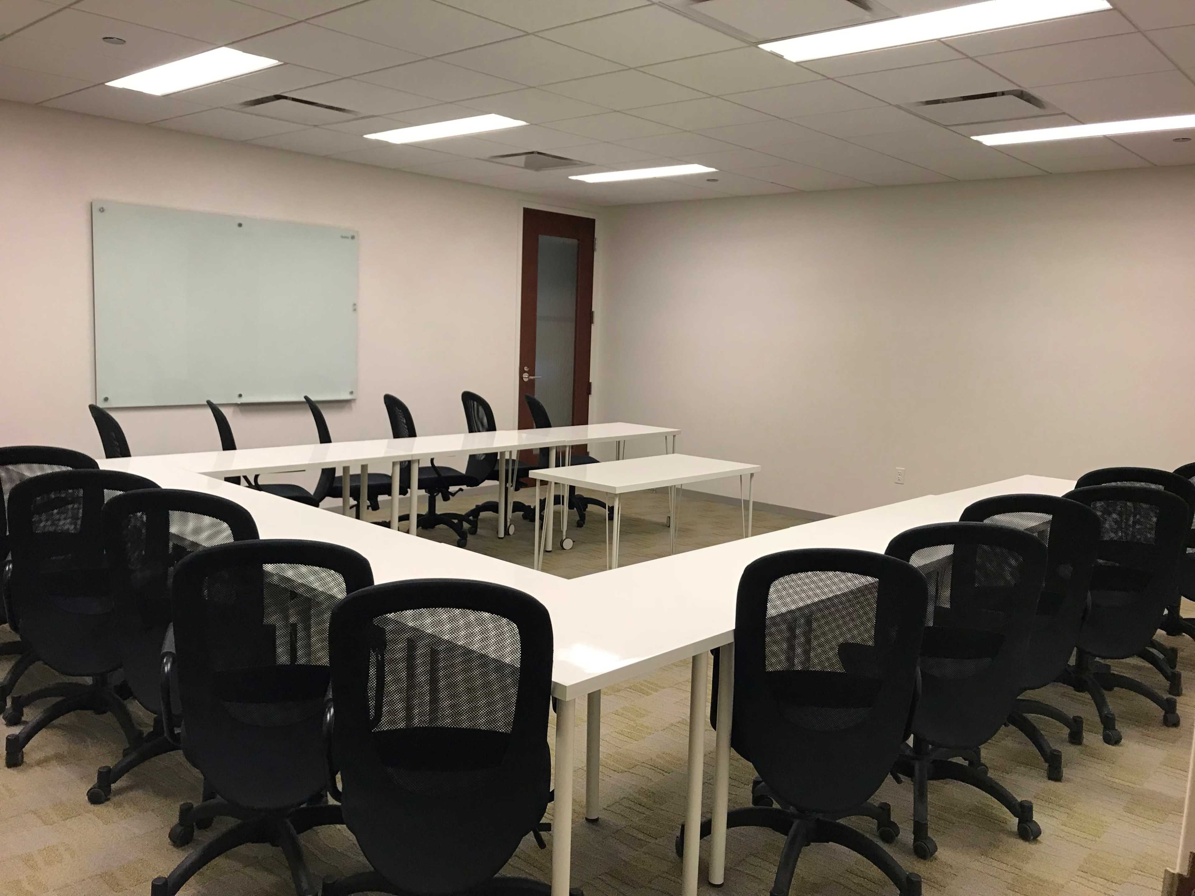 A conference room features a U-shaped arrangement of white tables surrounded by black office chairs, with a whiteboard on one wall.