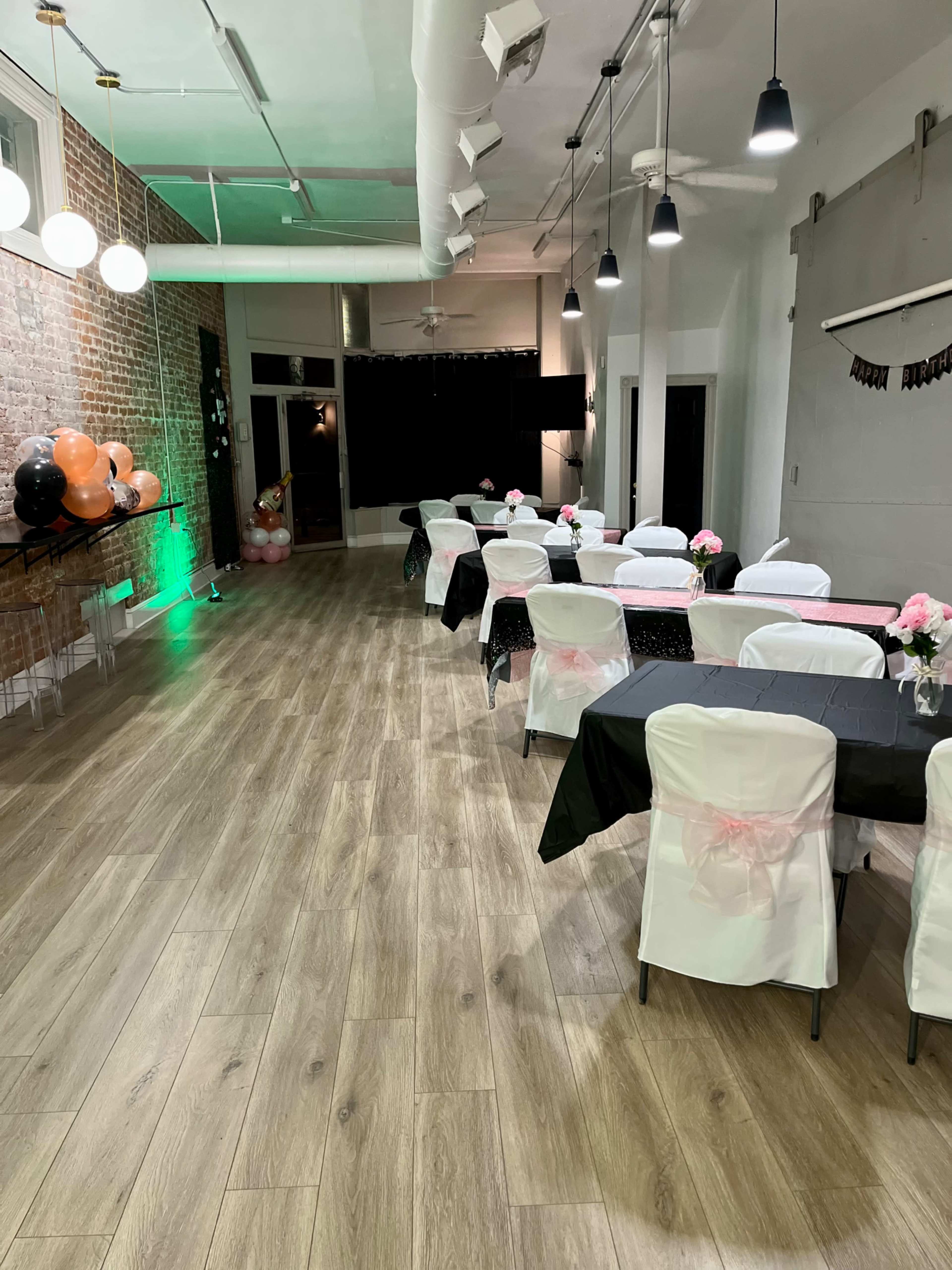 A decorated event space with tables set in a row, featuring black and white tablecloths and floral centerpieces, alongside balloons and dim lighting.