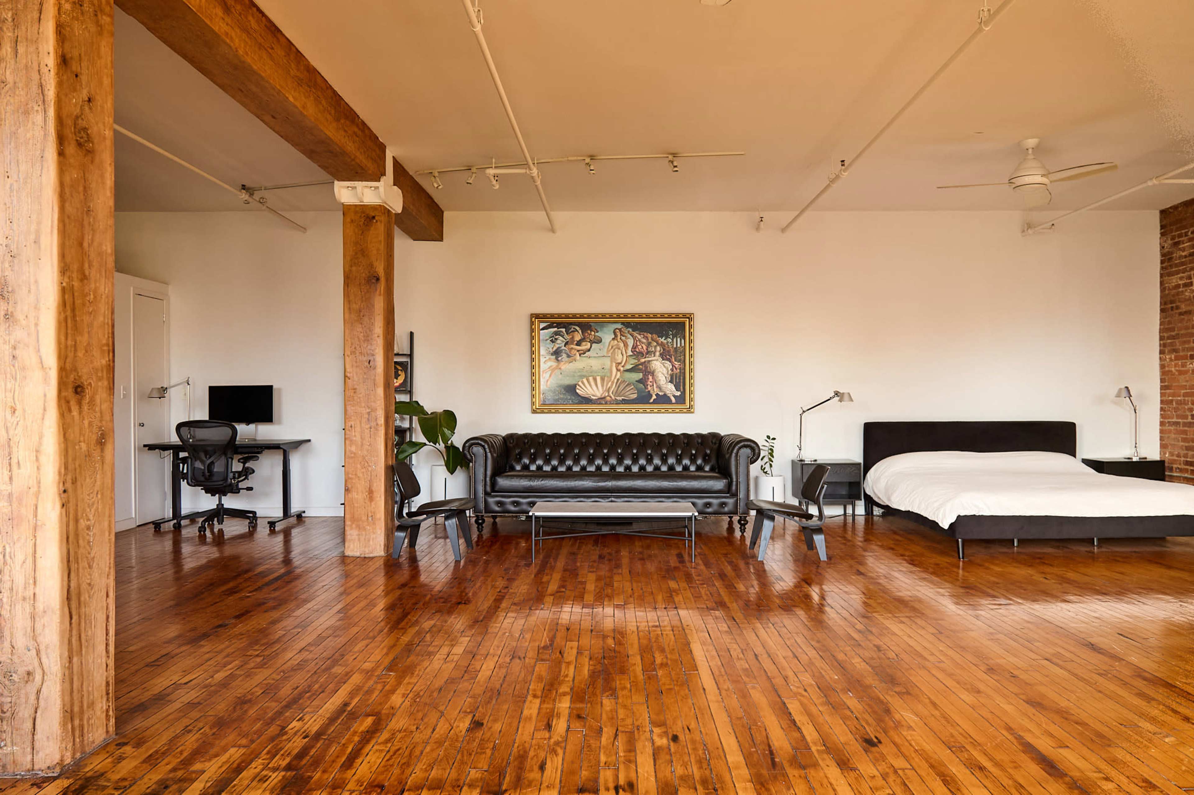 The image shows a spacious loft-style room featuring wooden flooring, a black leather sofa, a white bed, a desk with a chair, and a large artwork on the wall.