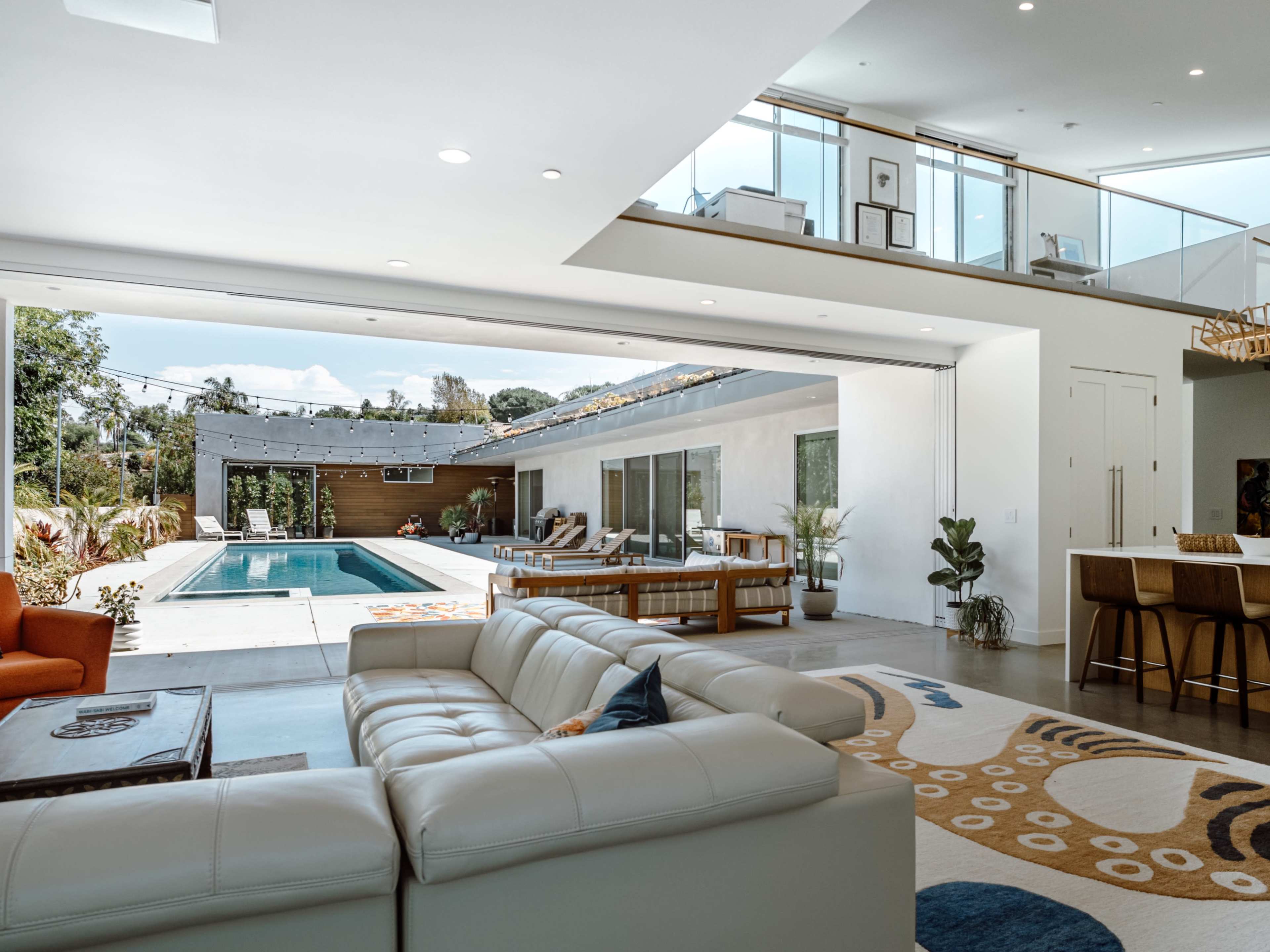 The image shows a modern, open-concept living area with a view of a pool, outdoor seating, and a dining space.