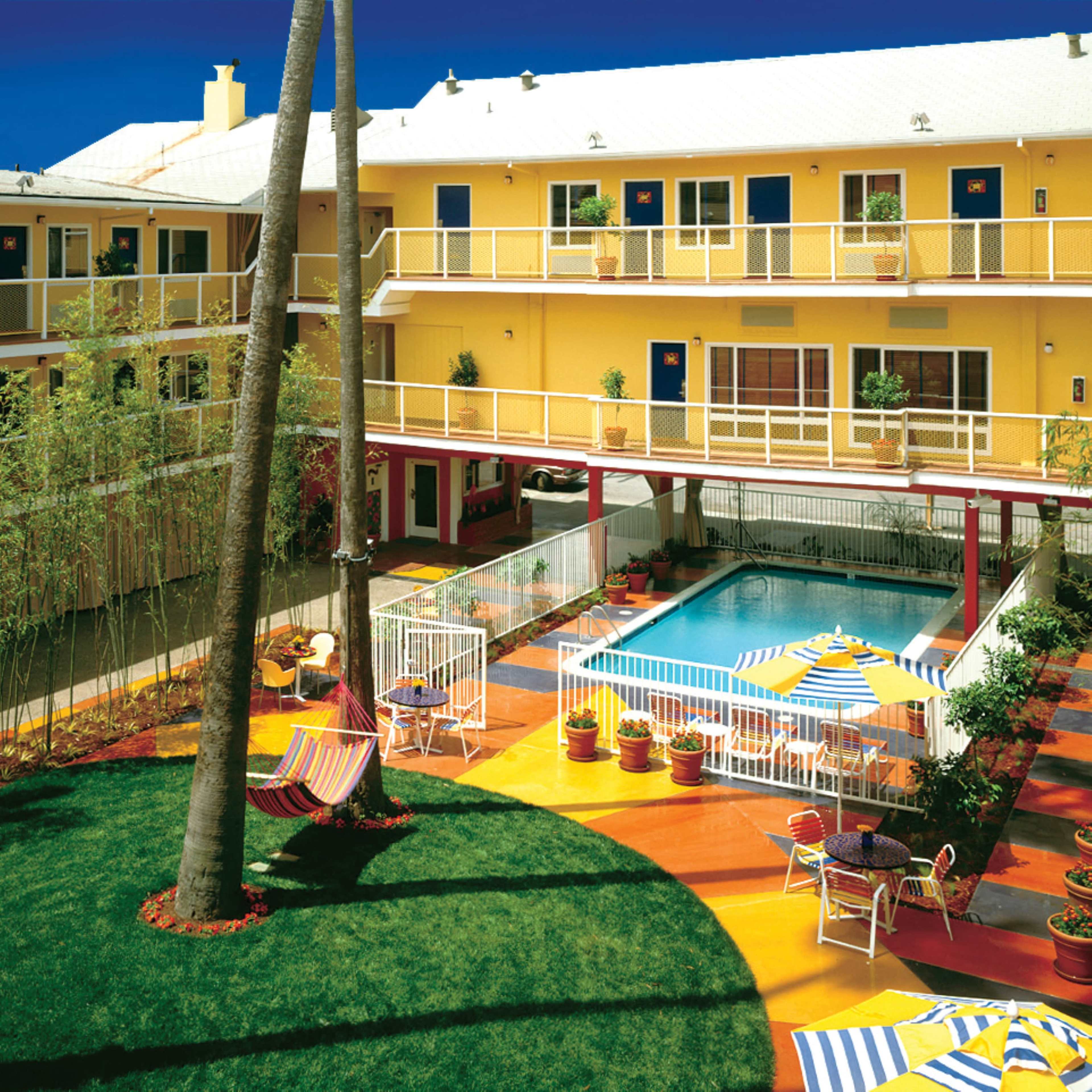 A colorful courtyard with a swimming pool surrounded by potted plants and lounge chairs, flanked by a three-story yellow building.