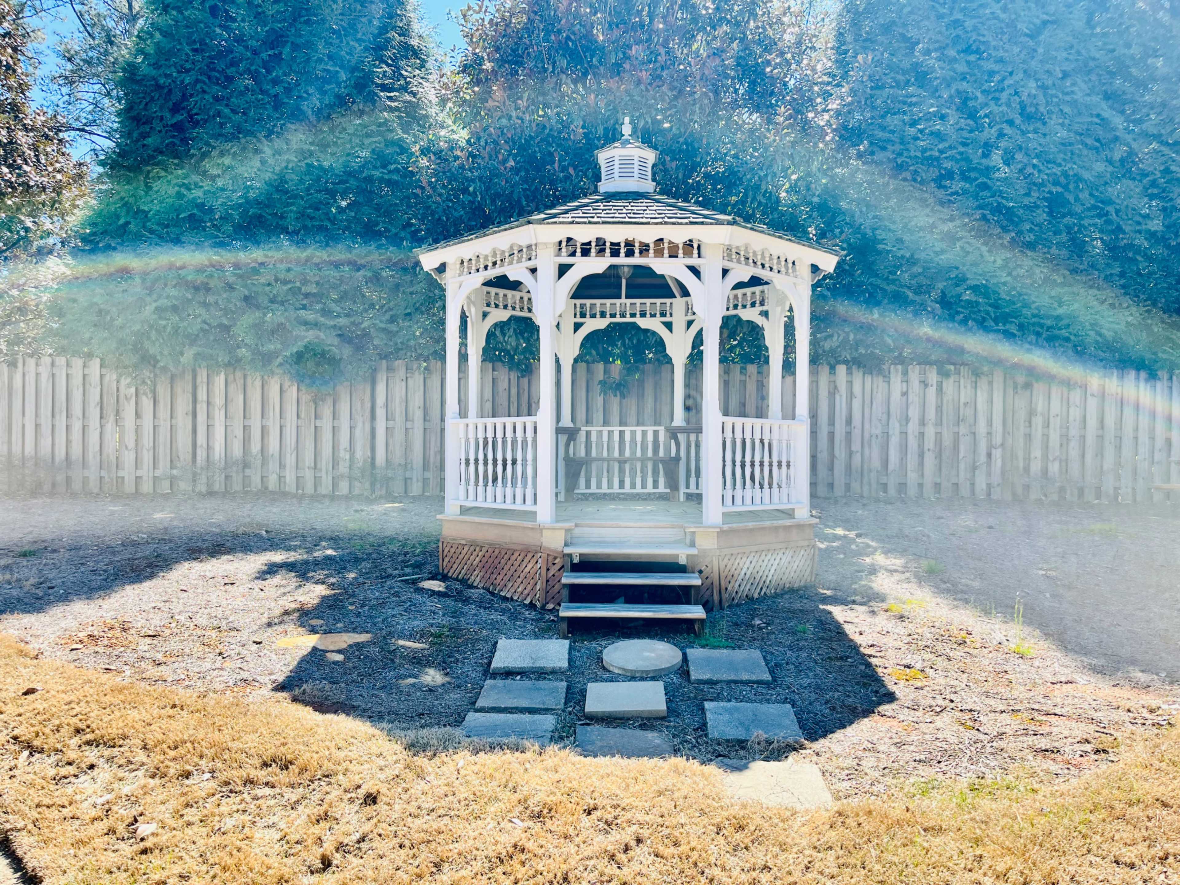A white gazebo with a shingled roof is positioned on a stone pathway in a grassy yard, surrounded by tall trees and a wooden fence.