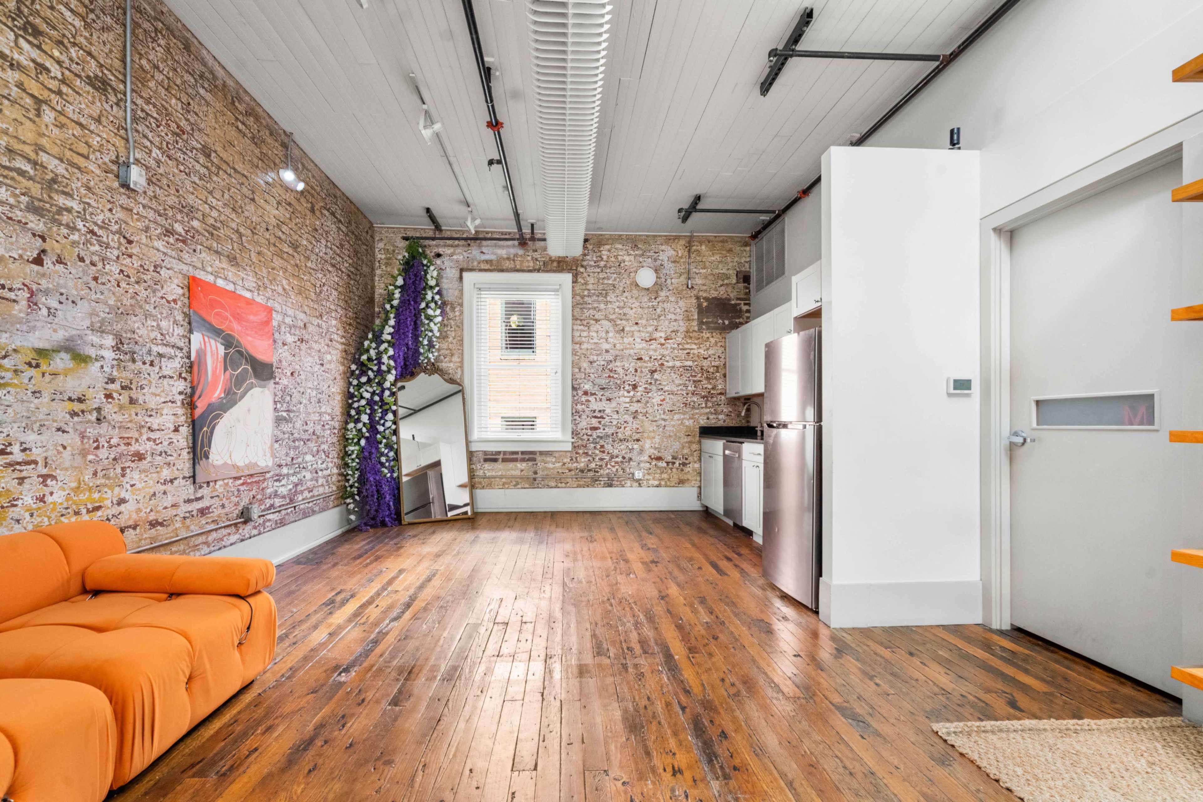 The image shows an open living space with exposed brick walls, wooden floors, a large orange sofa, and a kitchenette in the background.
