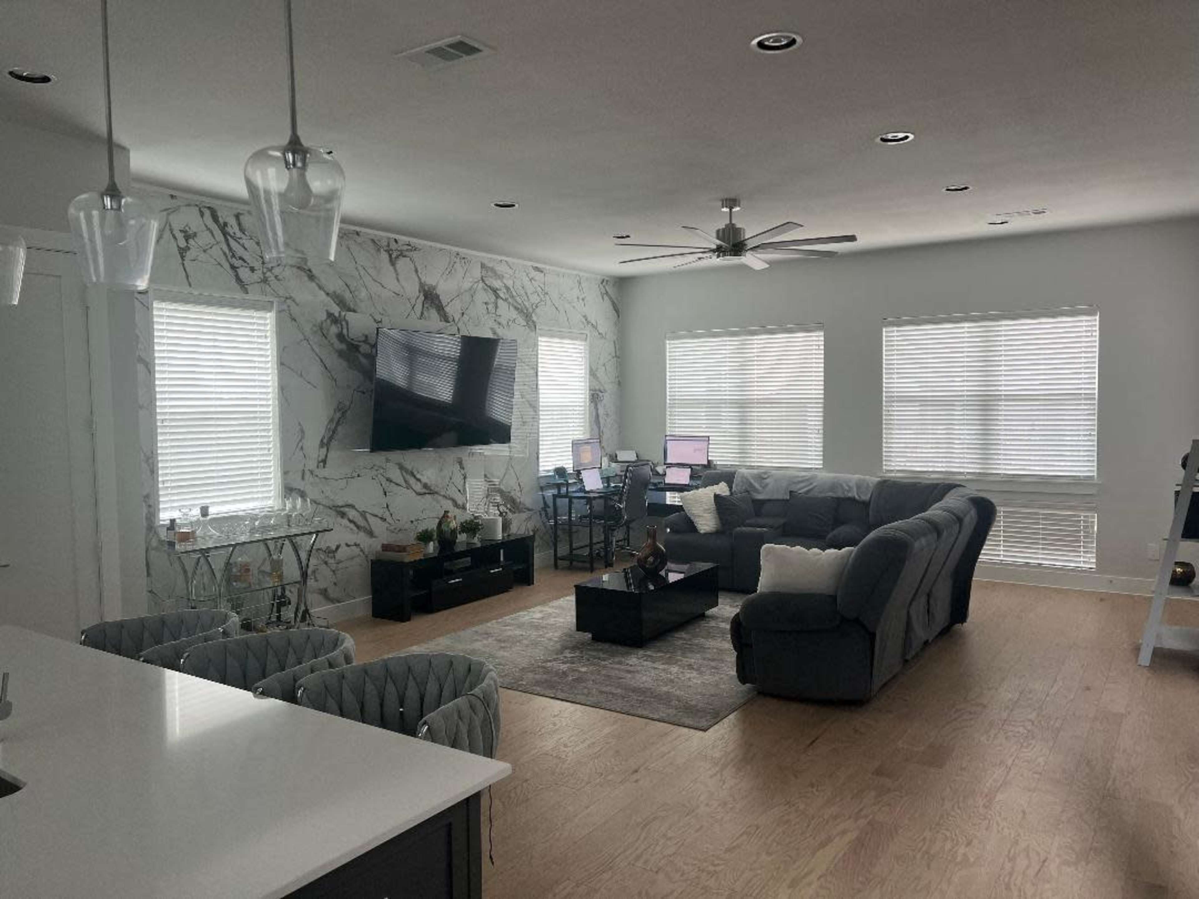 A modern living room with a sectional sofa, a coffee table, a wall-mounted TV, and two workstations against a marble accent wall.