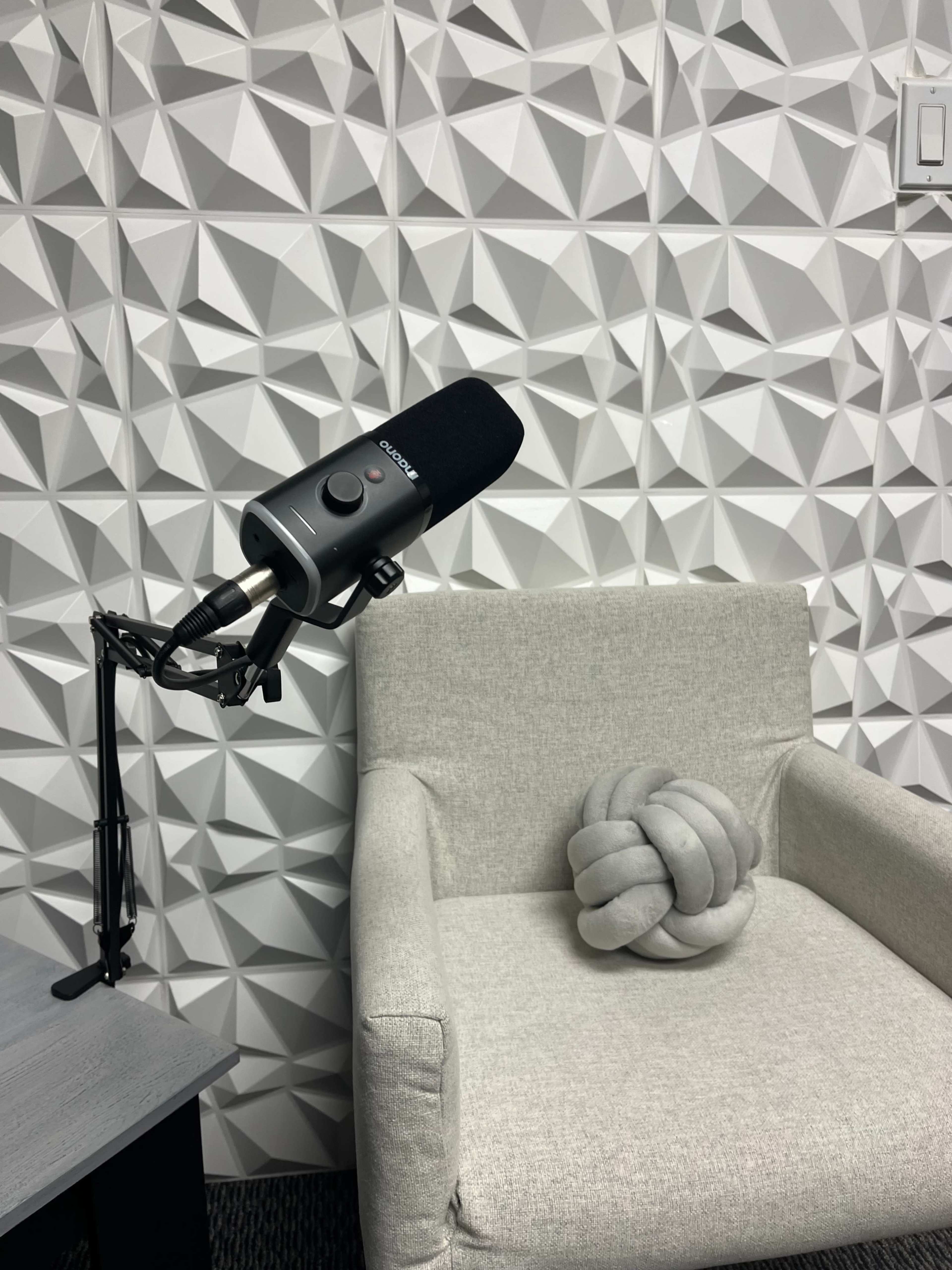 A microphone is mounted on an adjustable arm next to a light gray chair and a decorative knot-shaped pillow, set against a textured, three-dimensional wall.