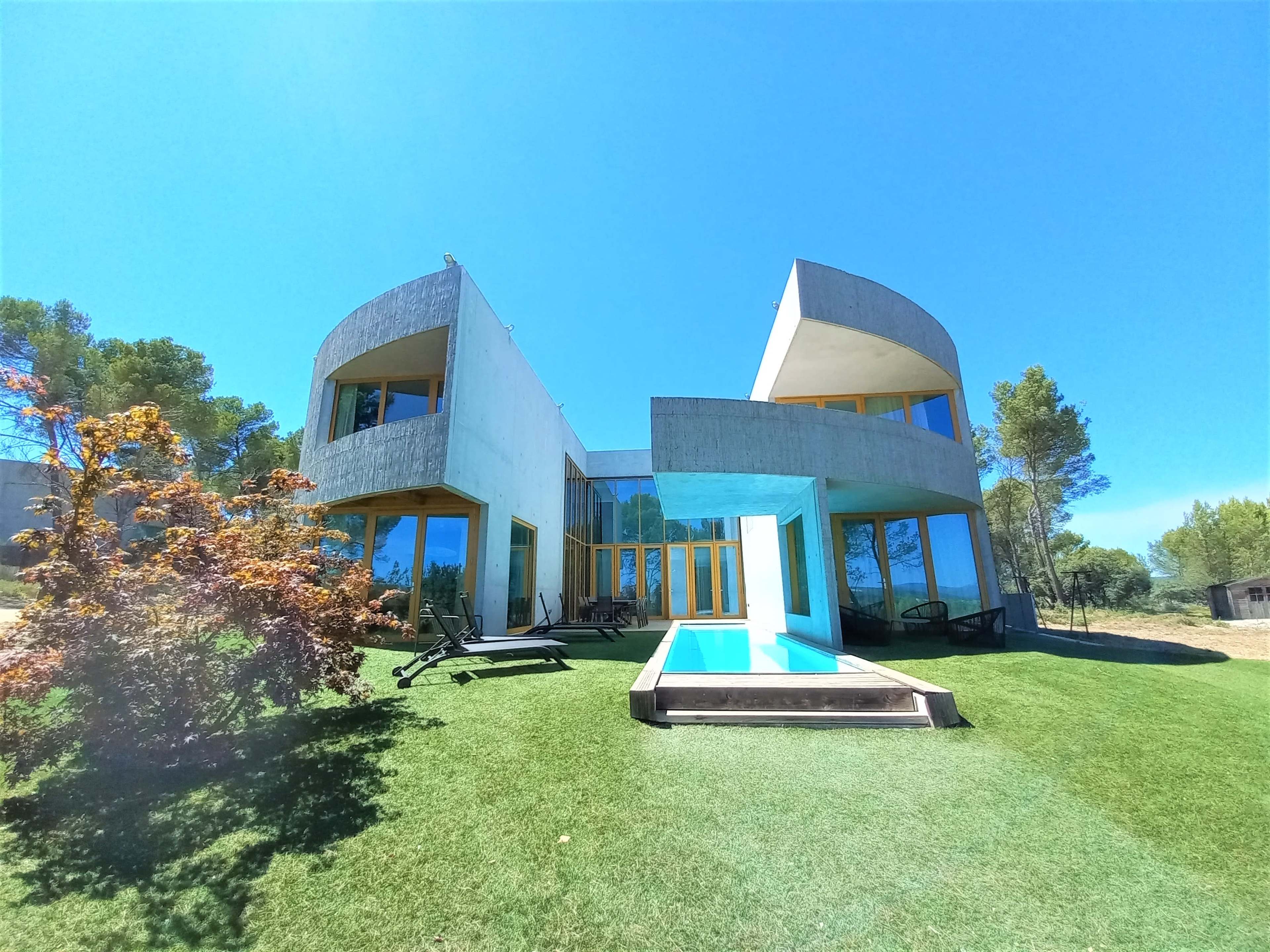 A modern house with large windows and a pool, situated on a grassy area surrounded by trees.
