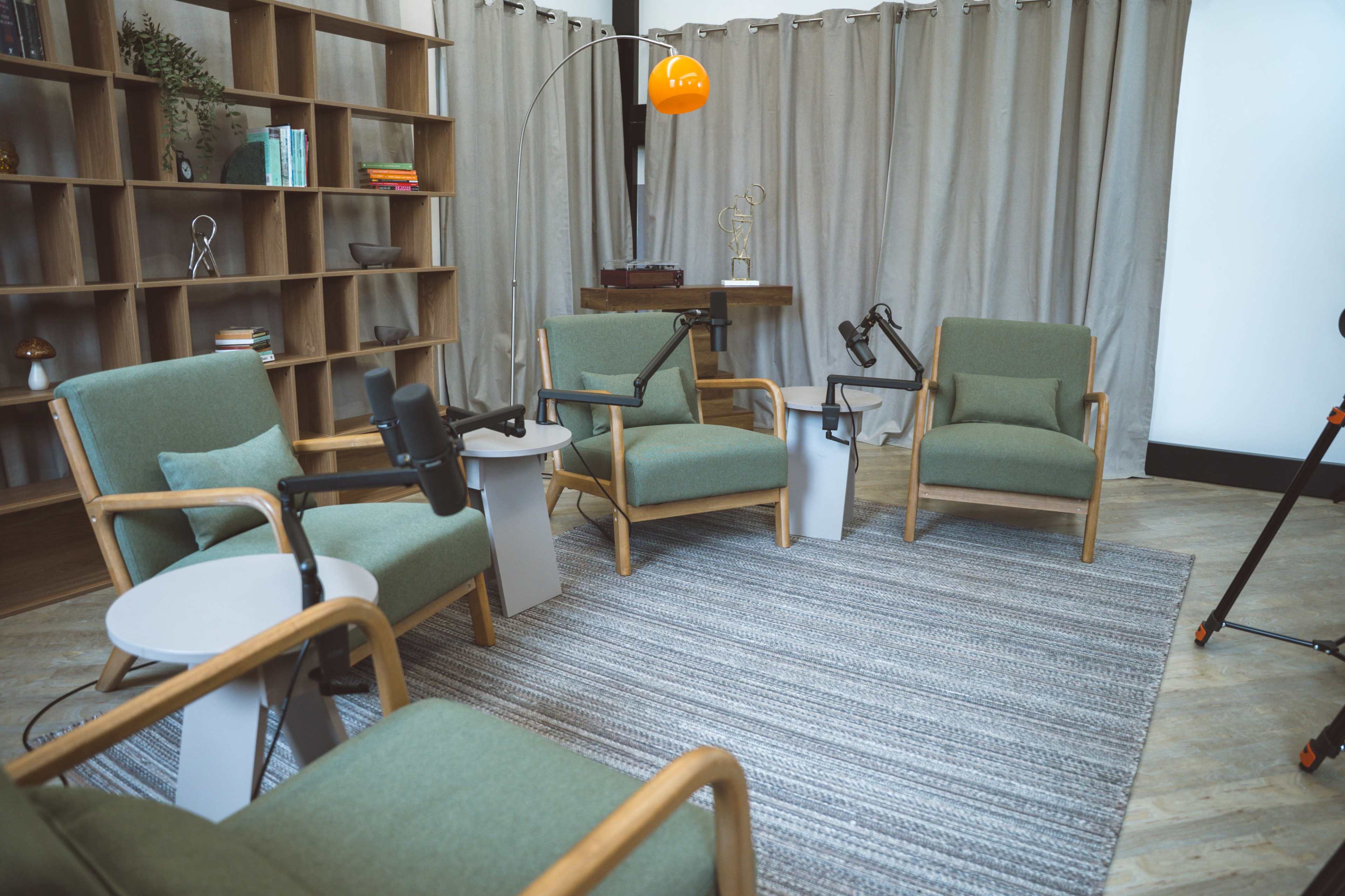 The image shows a cozy interview setting featuring four green armchairs arranged around two small tables, with microphones positioned on stands and a curtain backdrop.