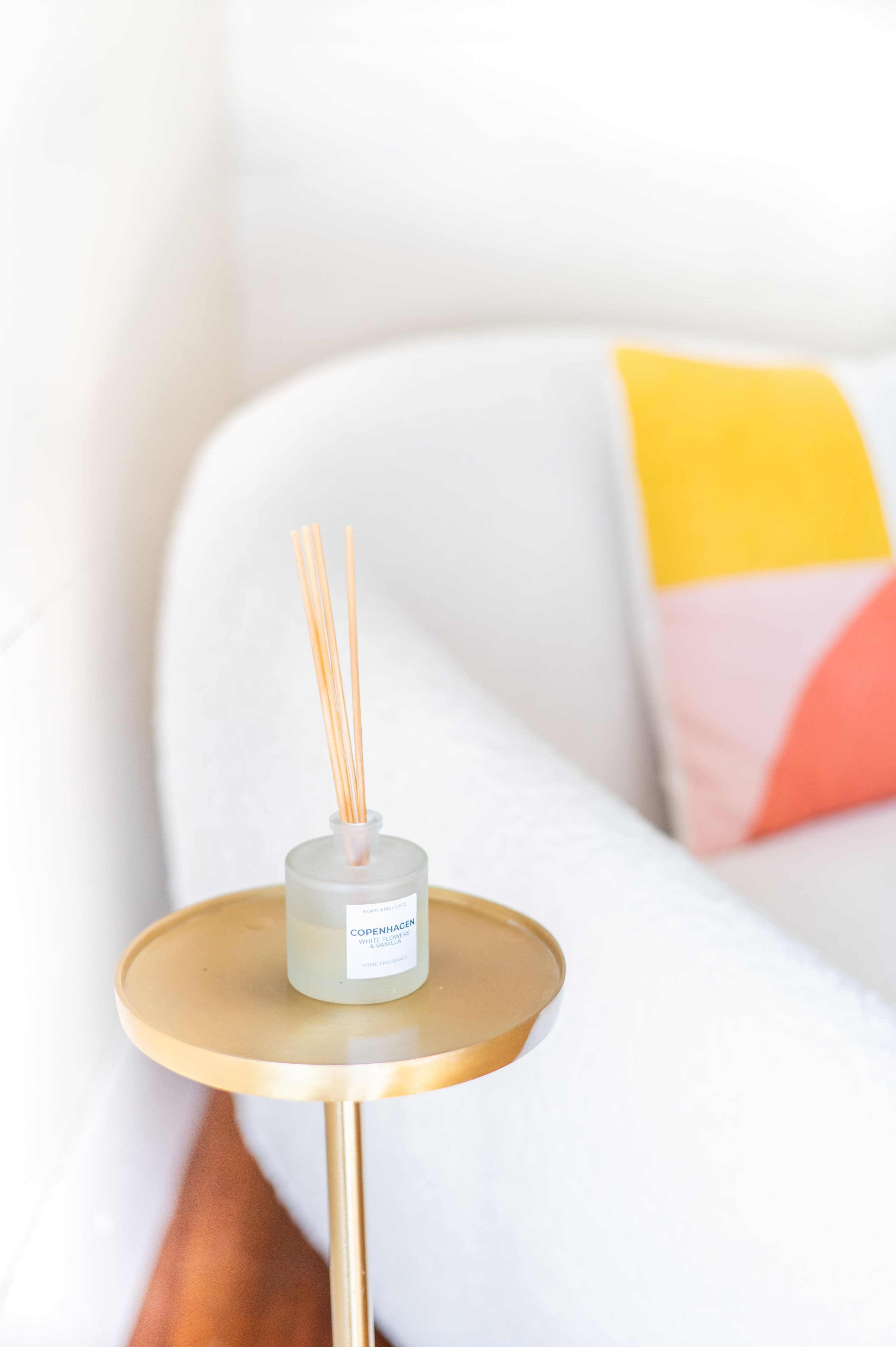 A round, gold side table holds a white candle with reed diffusers next to a pale, textured couch featuring colorful cushions.
