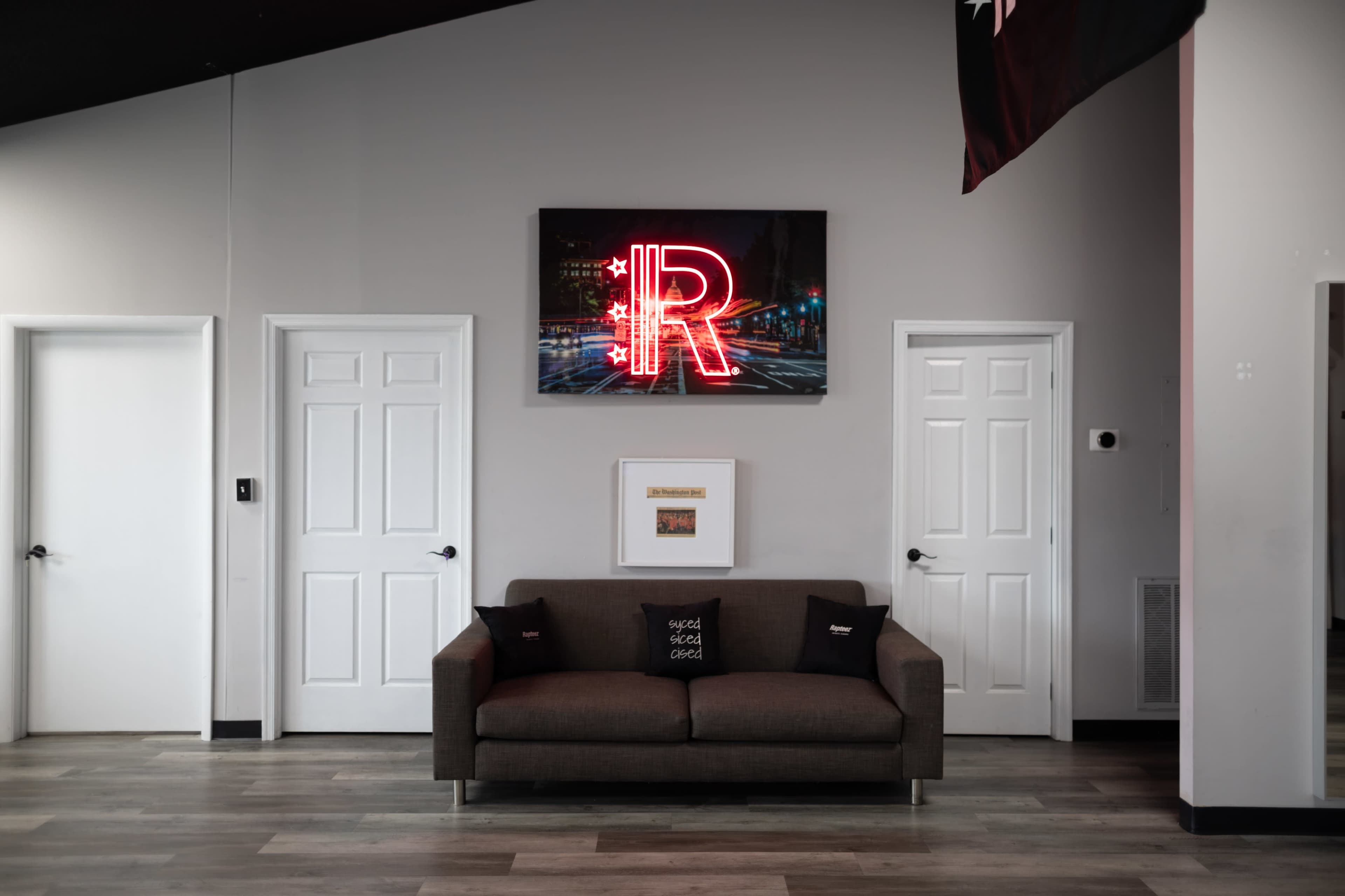 A modern waiting area features a brown sofa with decorative pillows, two white doors, and a neon sign with the letter "R" mounted on the wall above.