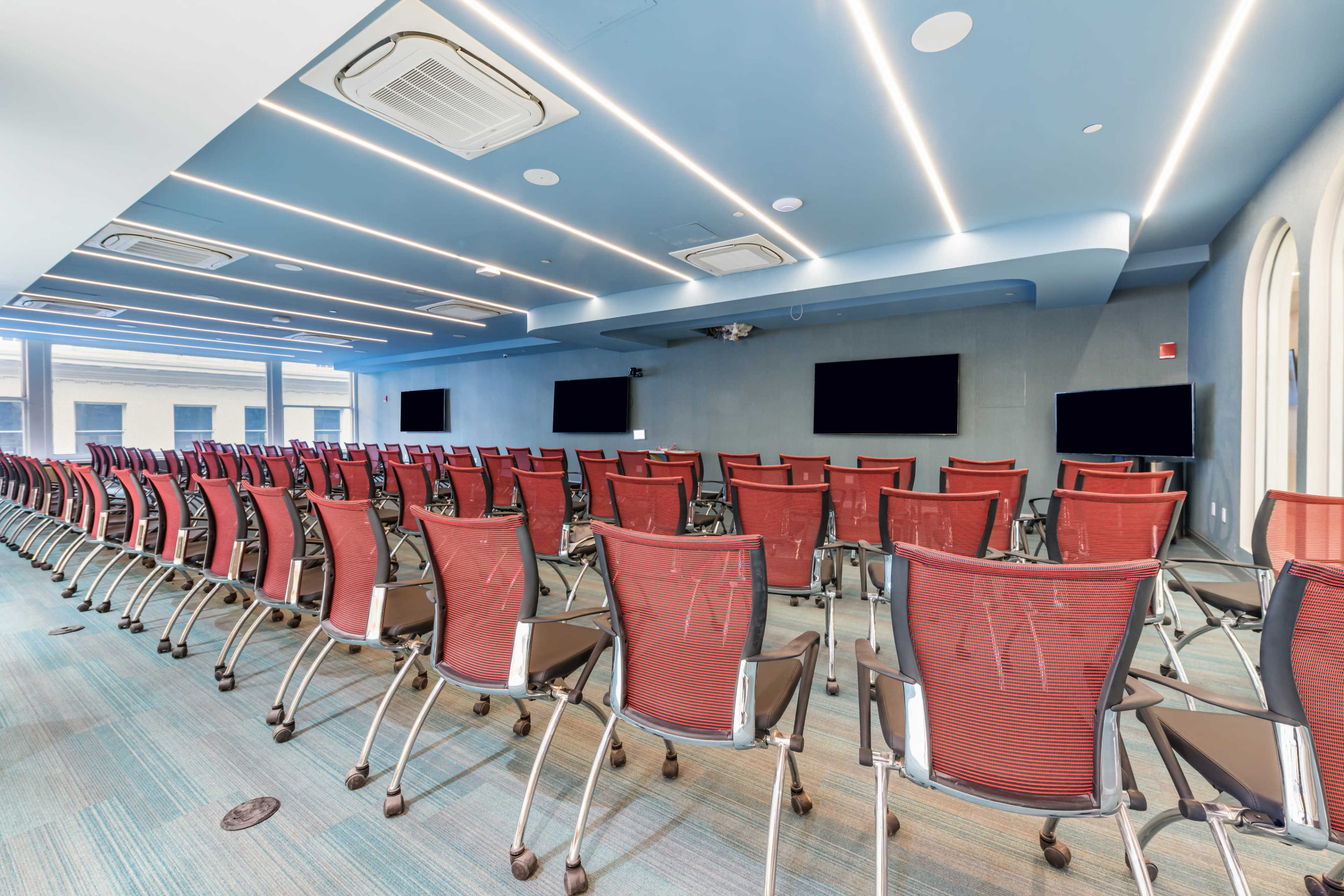 The image shows a modern conference room with rows of red mesh chairs facing two large screens on a gray wall.