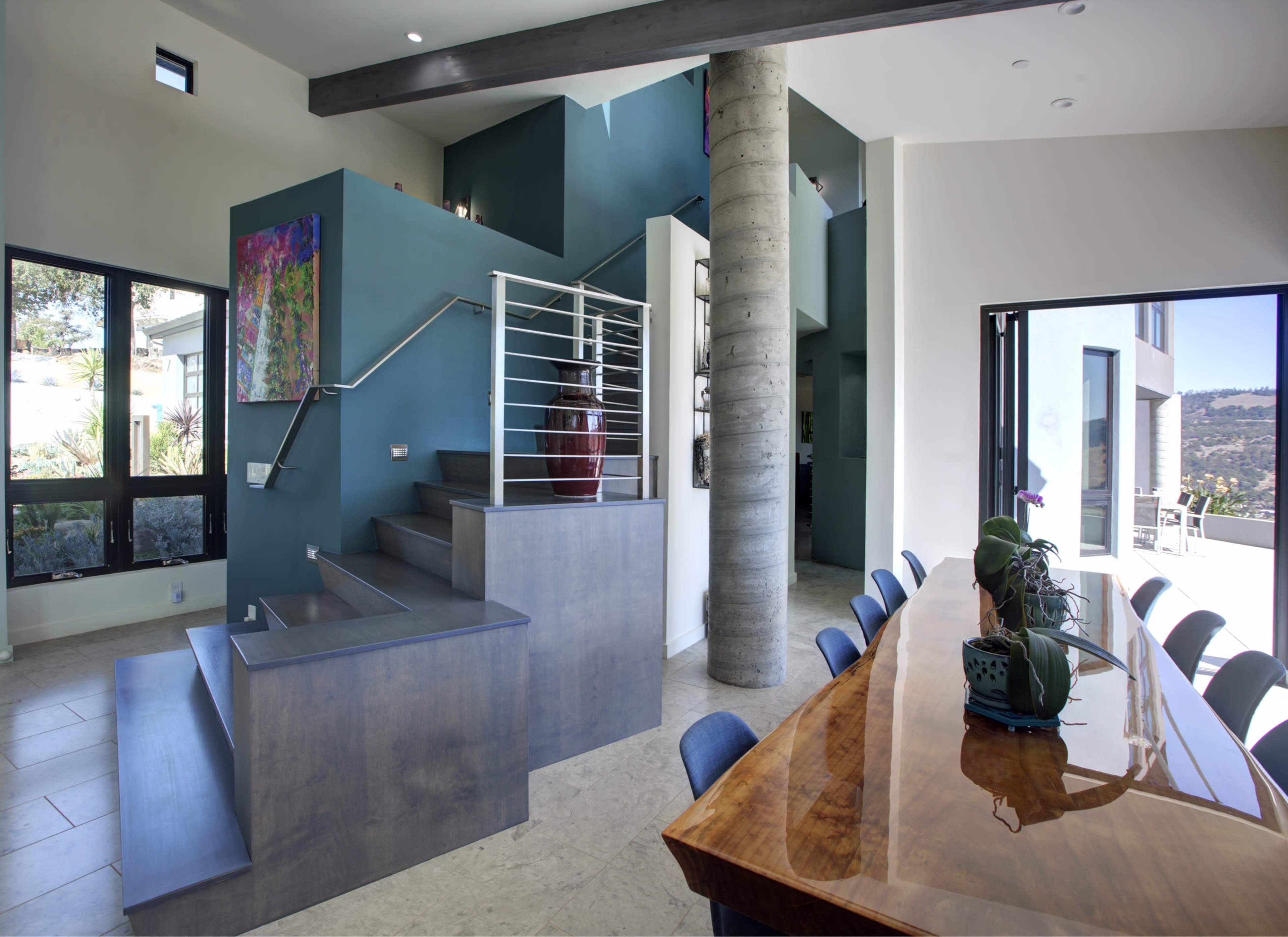 A modern interior featuring a staircase with a sleek railing and a long dining table beside large windows that overlook a scenic view.
