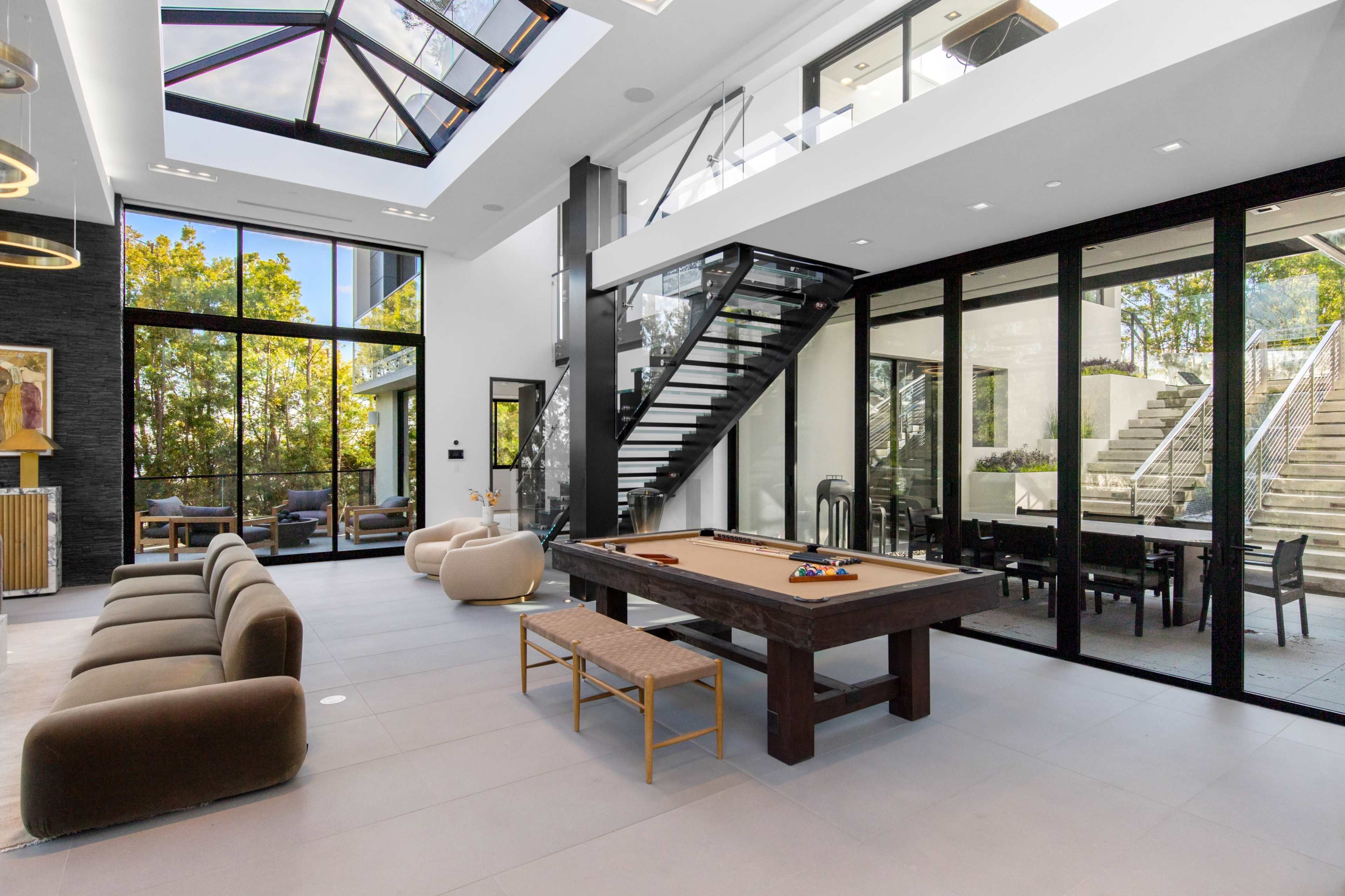 The image shows a modern interior space featuring a game table, a staircase, large glass walls, and a view of greenery outside.