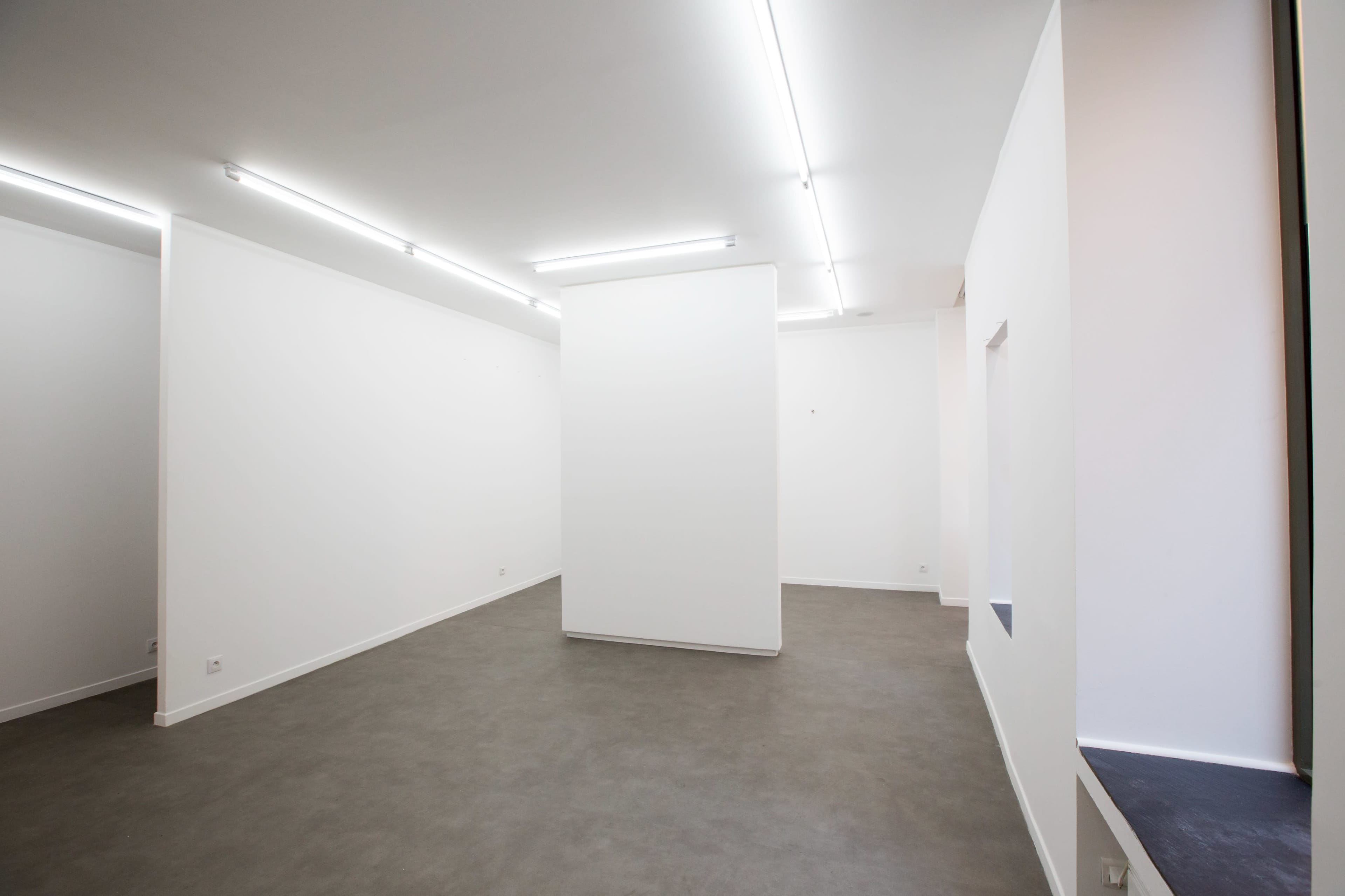 The image shows a spacious, minimally decorated interior with white walls and a gray floor, featuring a central white column.