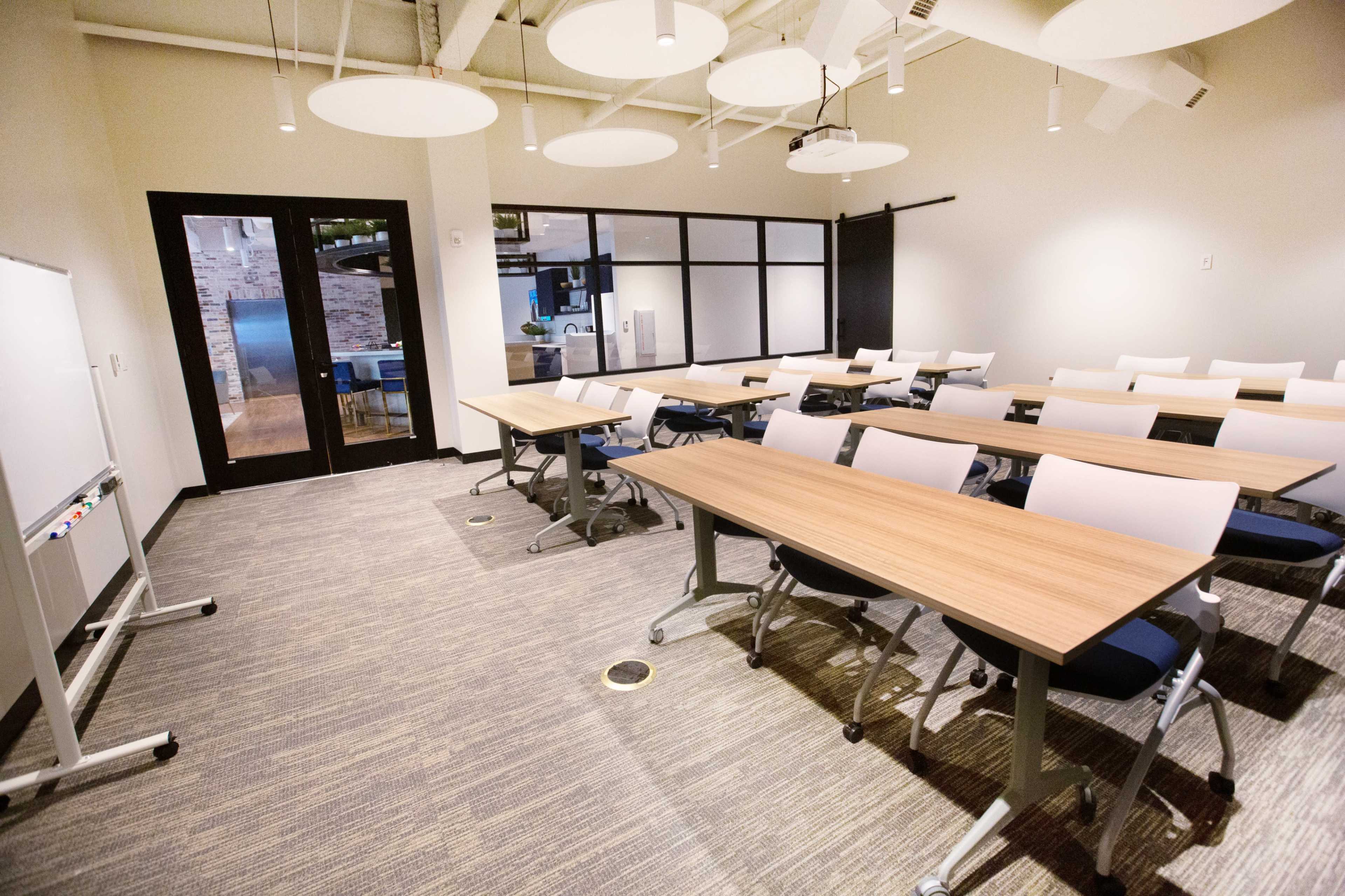A classroom setup with several rectangular tables and chairs arranged in rows, a whiteboard, and large windows along one wall.