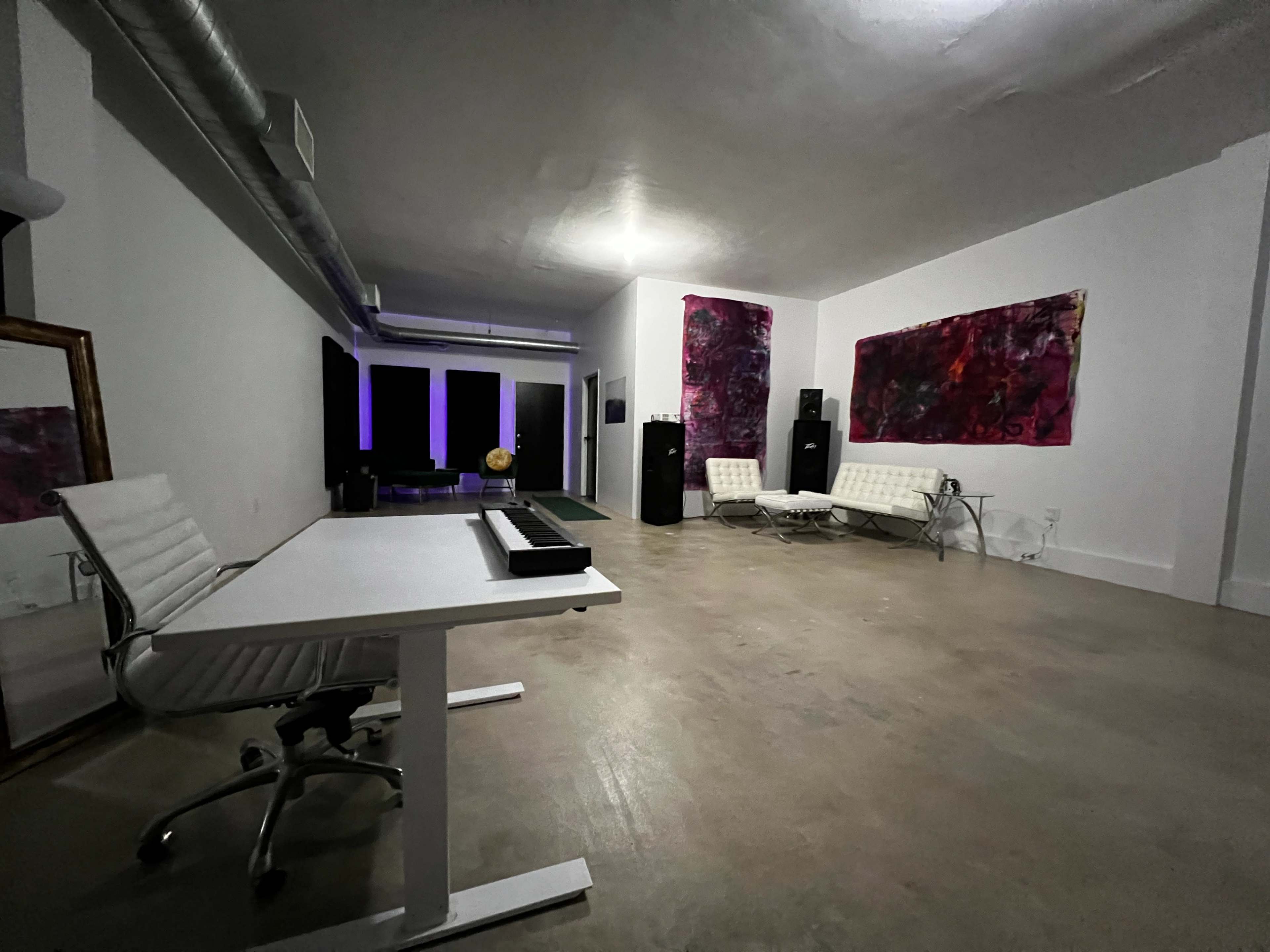 A spacious, minimalist room with concrete flooring, a white desk and chair, and two white lounge chairs near colorful abstract artwork on the walls.