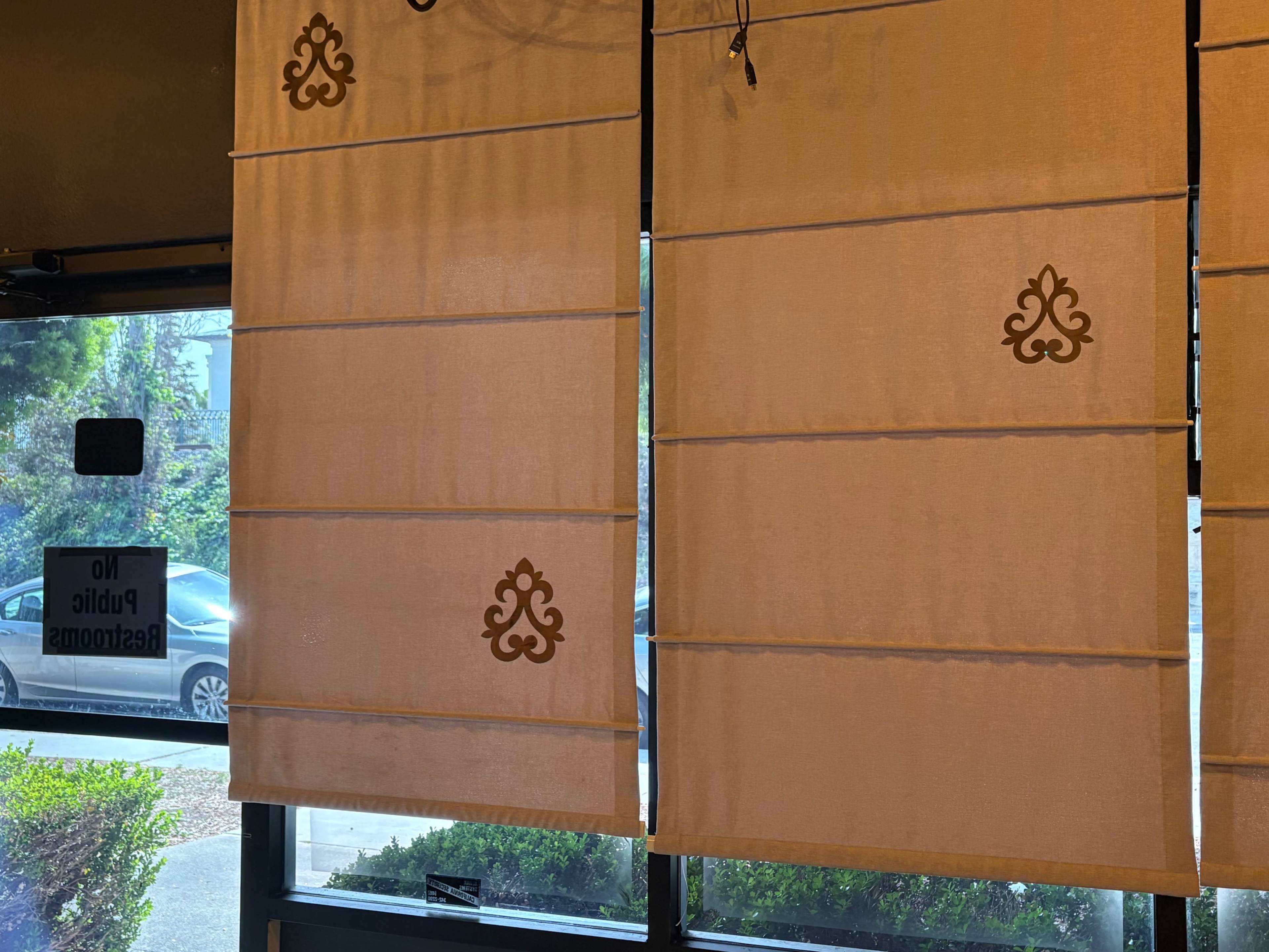 The image shows two partially opened fabric window shades with decorative patterns, set against a backdrop of a view outside.