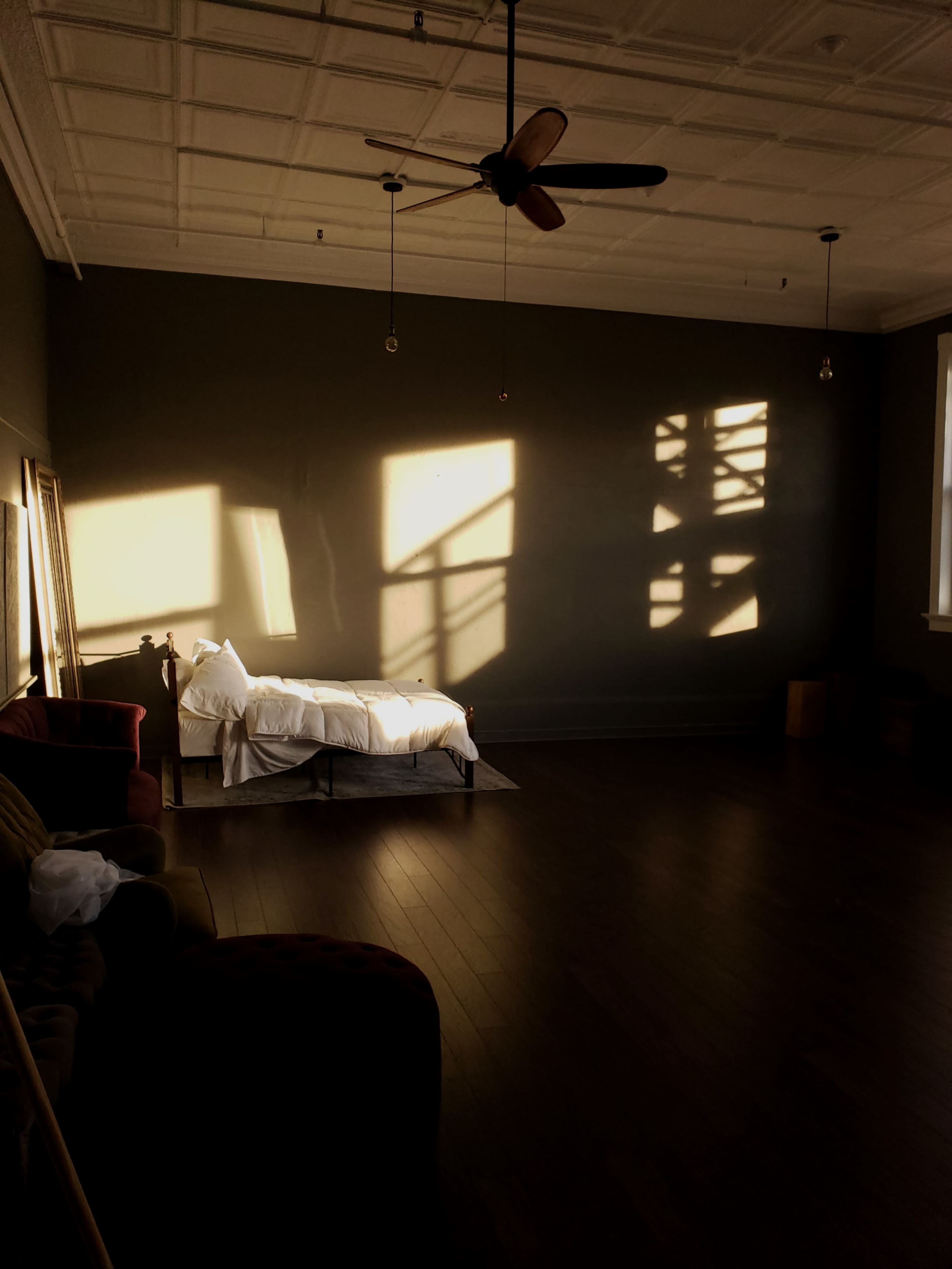 The room features a bed with white linens, a ceiling fan, and shadows of a window frame cast on the dark wall.
