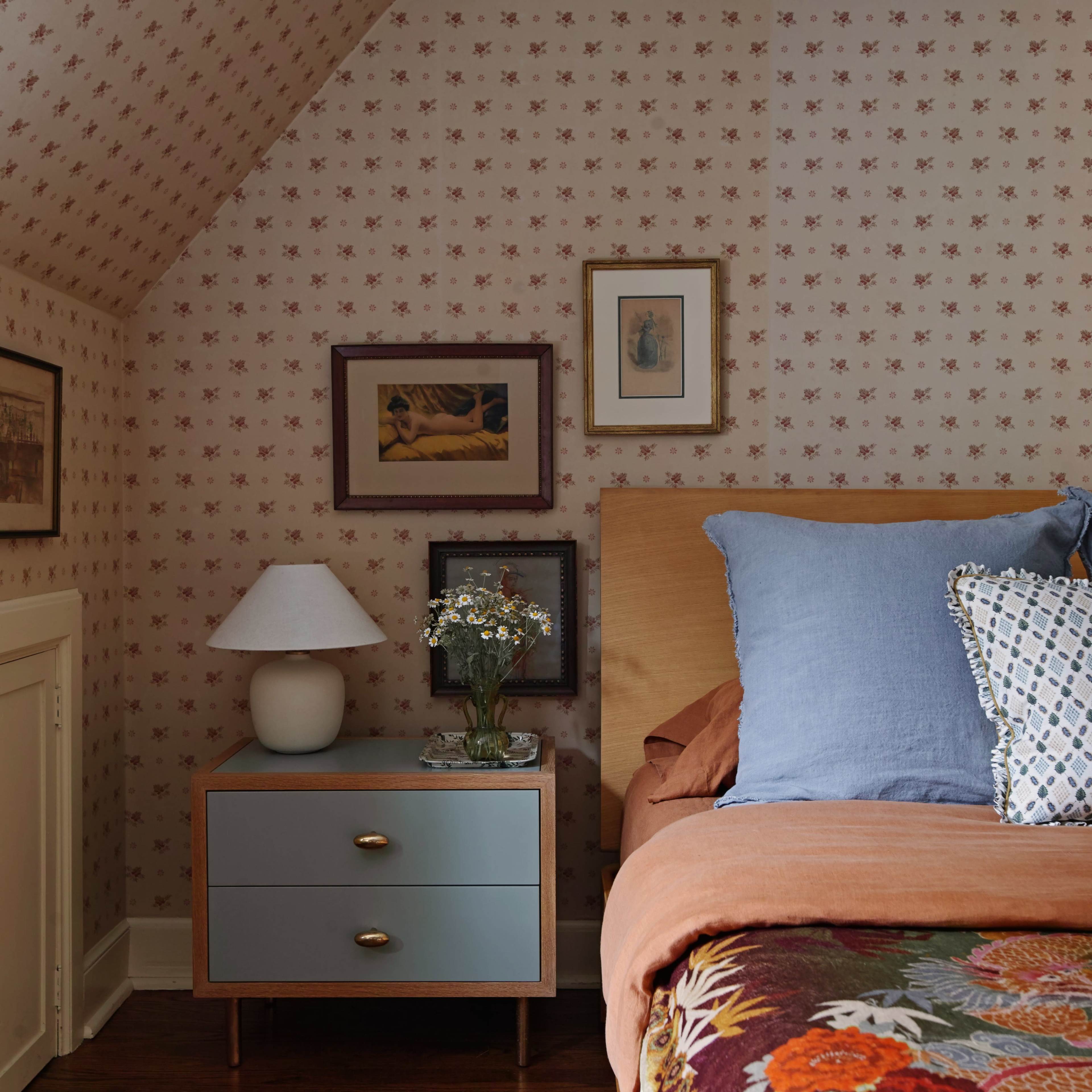 The image shows a cozy bedroom with a patterned wallpaper, a bed with colorful linens, a small nightstand, and framed artwork on the walls.