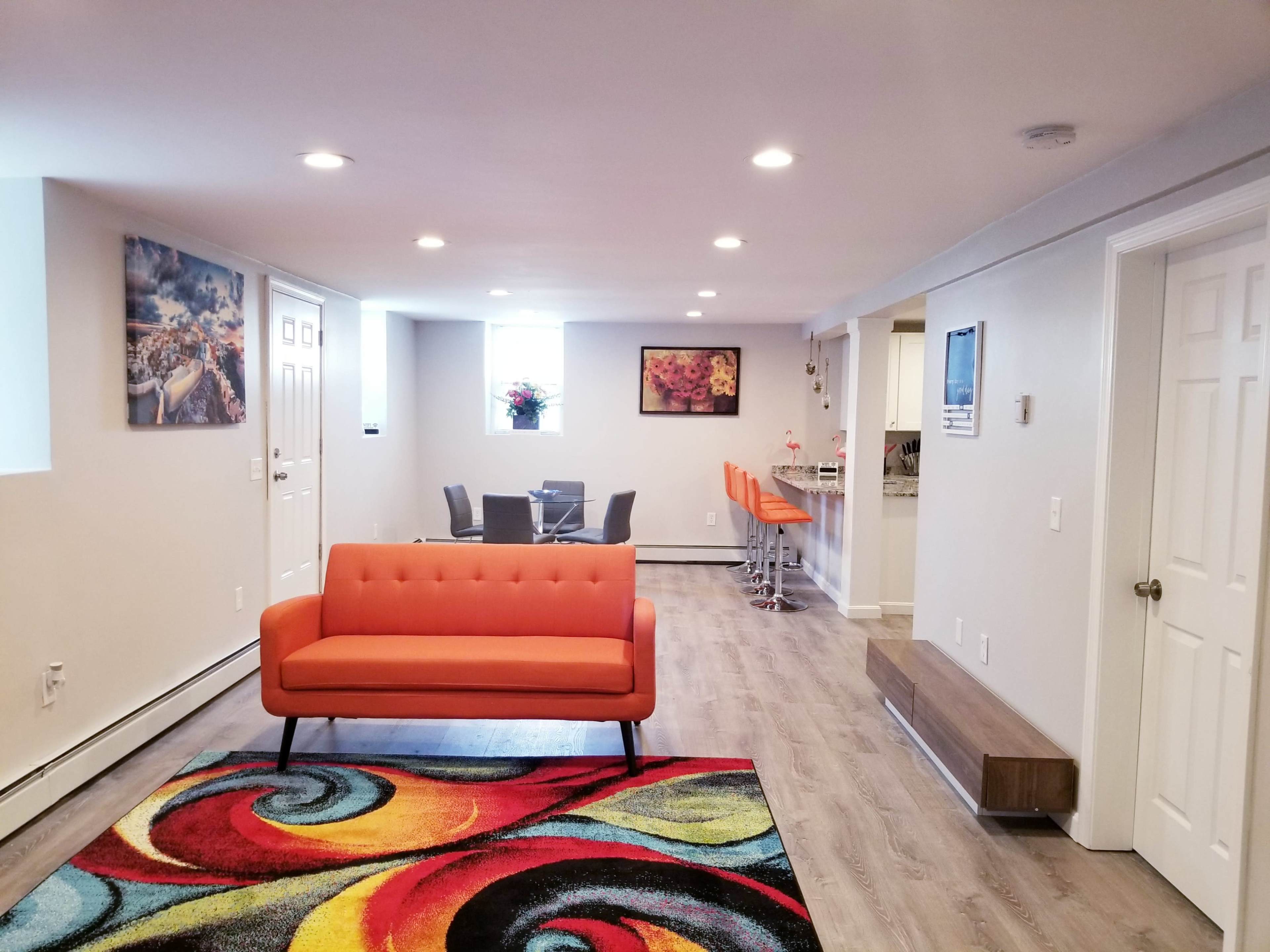 A brightly lit modern living room featuring an orange sofa, a colorful patterned rug, and a dining area with a table and chairs in the background.