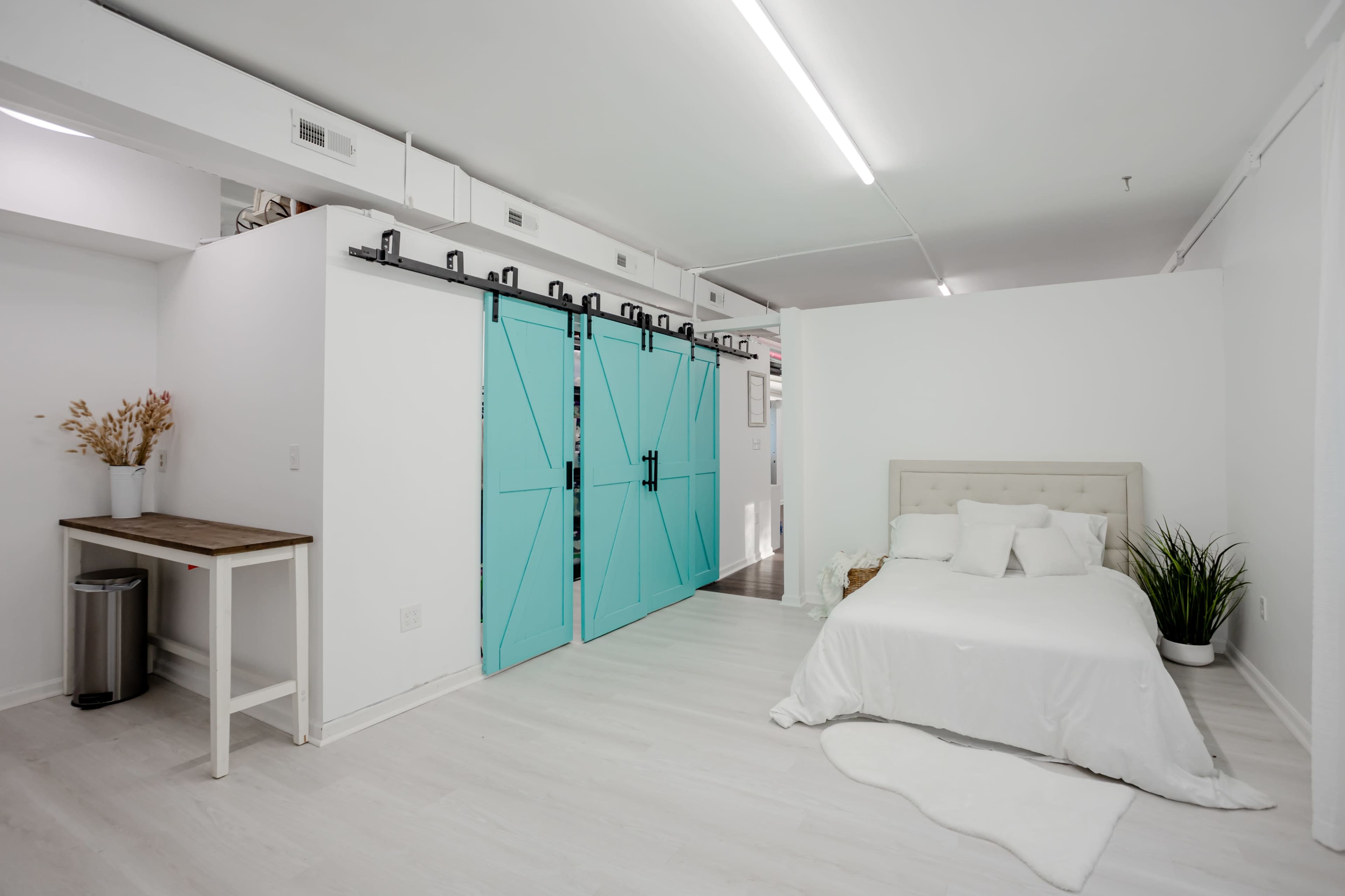 A minimalist room features a bed with white linens, a turquoise barn door, and a small table beside a trash can.