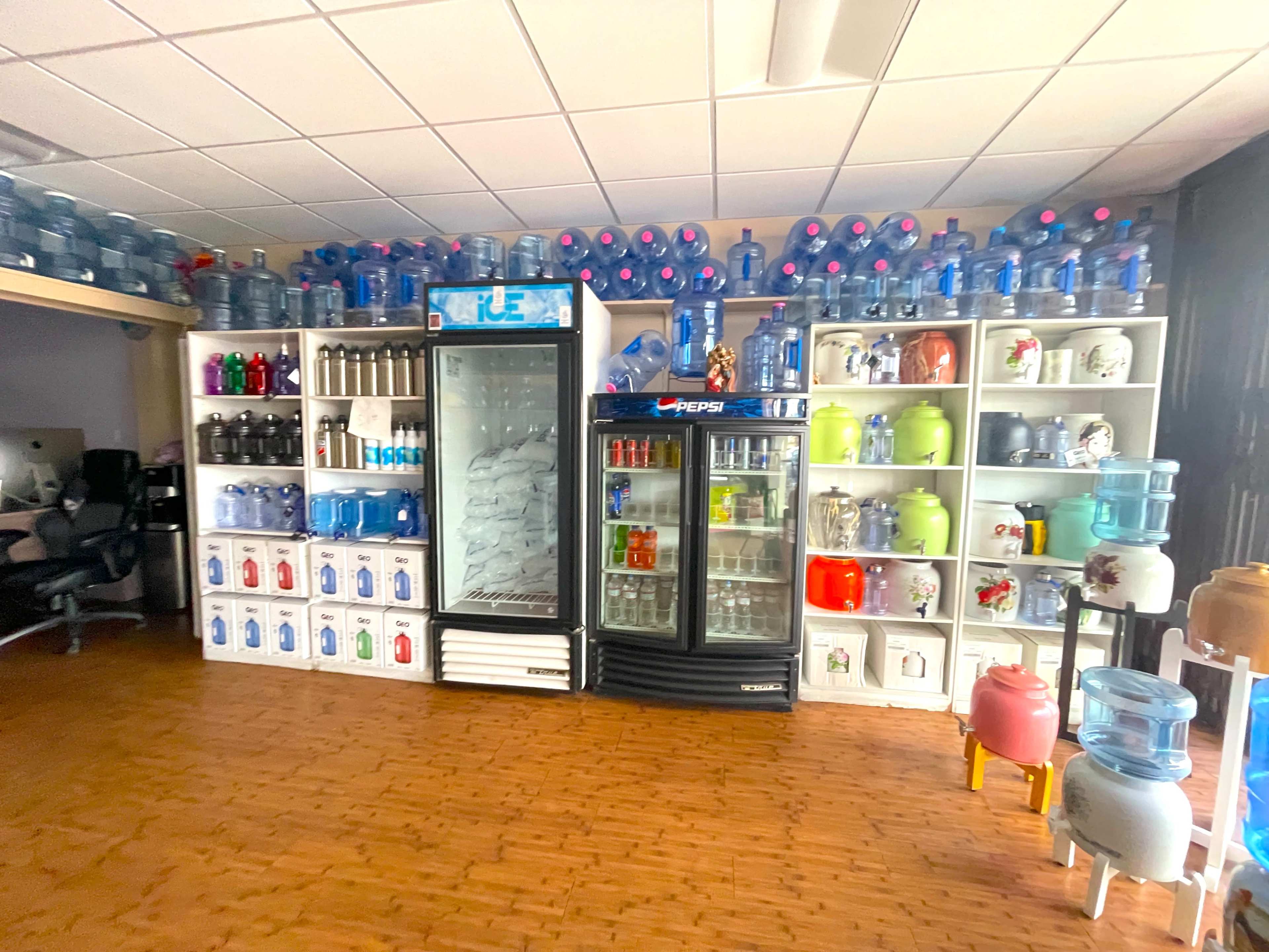 The image shows a room arranged with shelves filled with various types of water containers and dispensers, including a large refrigerator and several colored jugs.