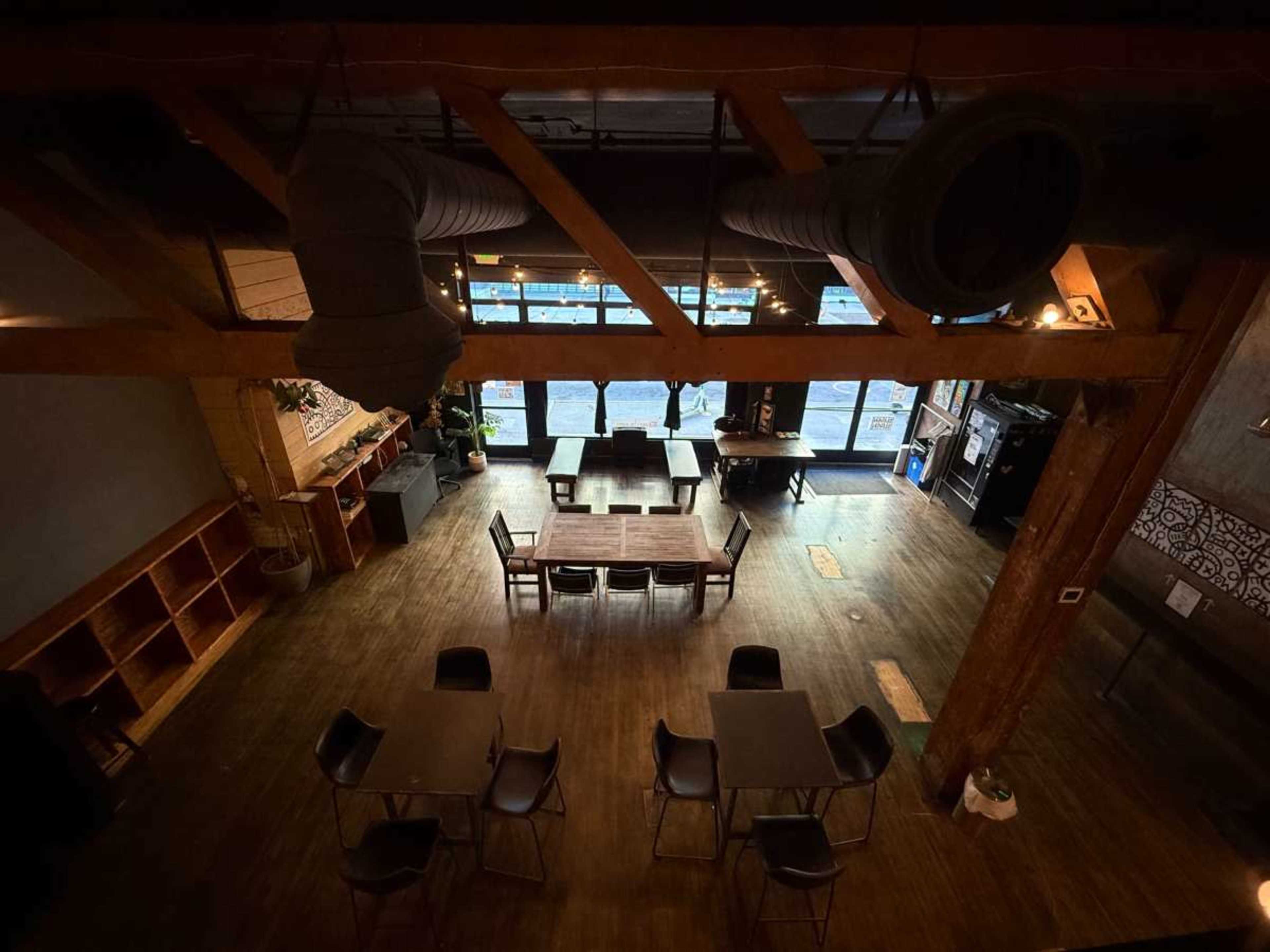 The image shows a spacious interior of a café or restaurant featuring wooden beams, tables, and a large window with a view outside.