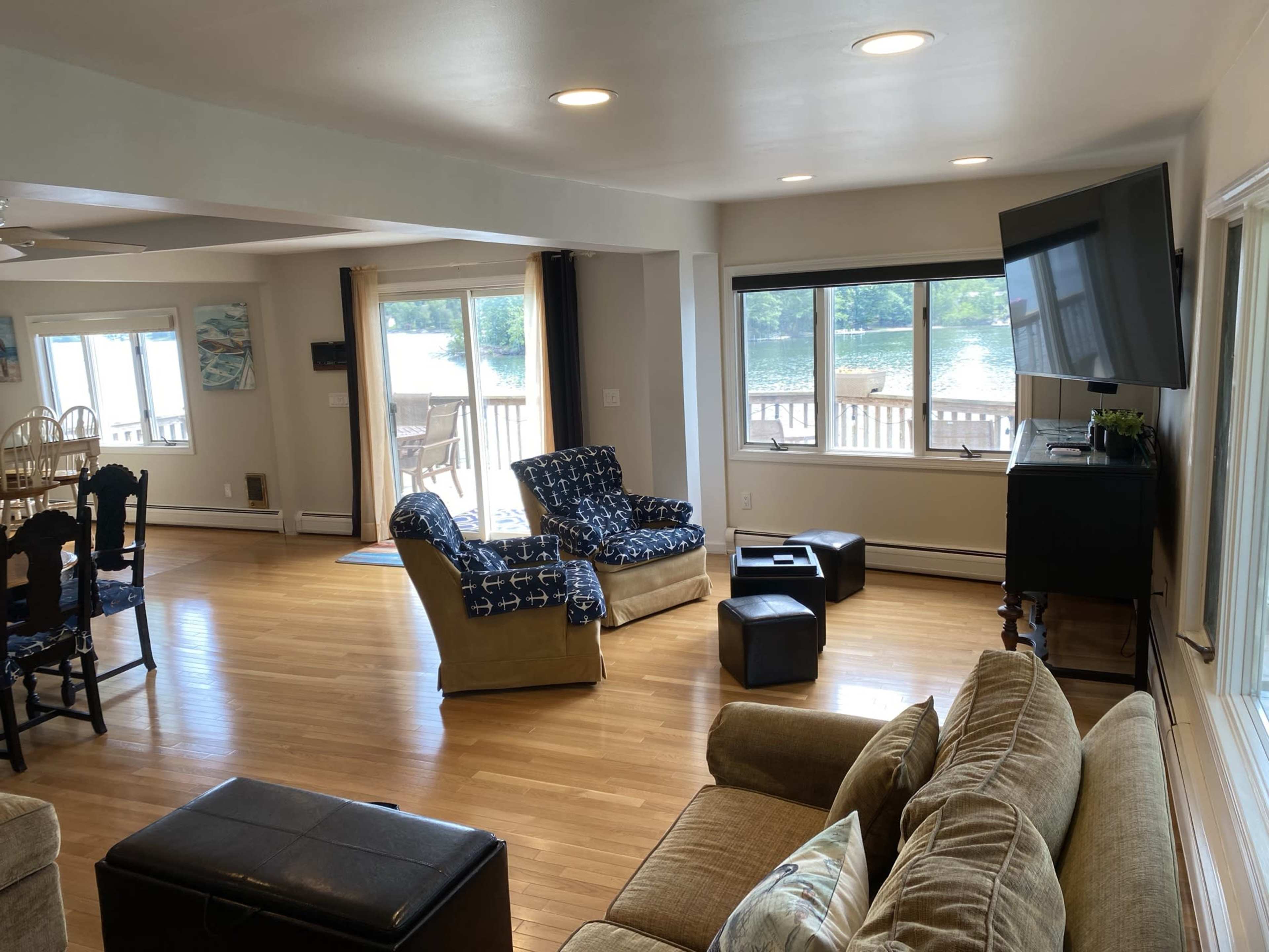 The image shows a spacious living area with wooden floors, two patterned chairs, a sofa, and a television mounted on the wall, alongside large windows allowing natural light.