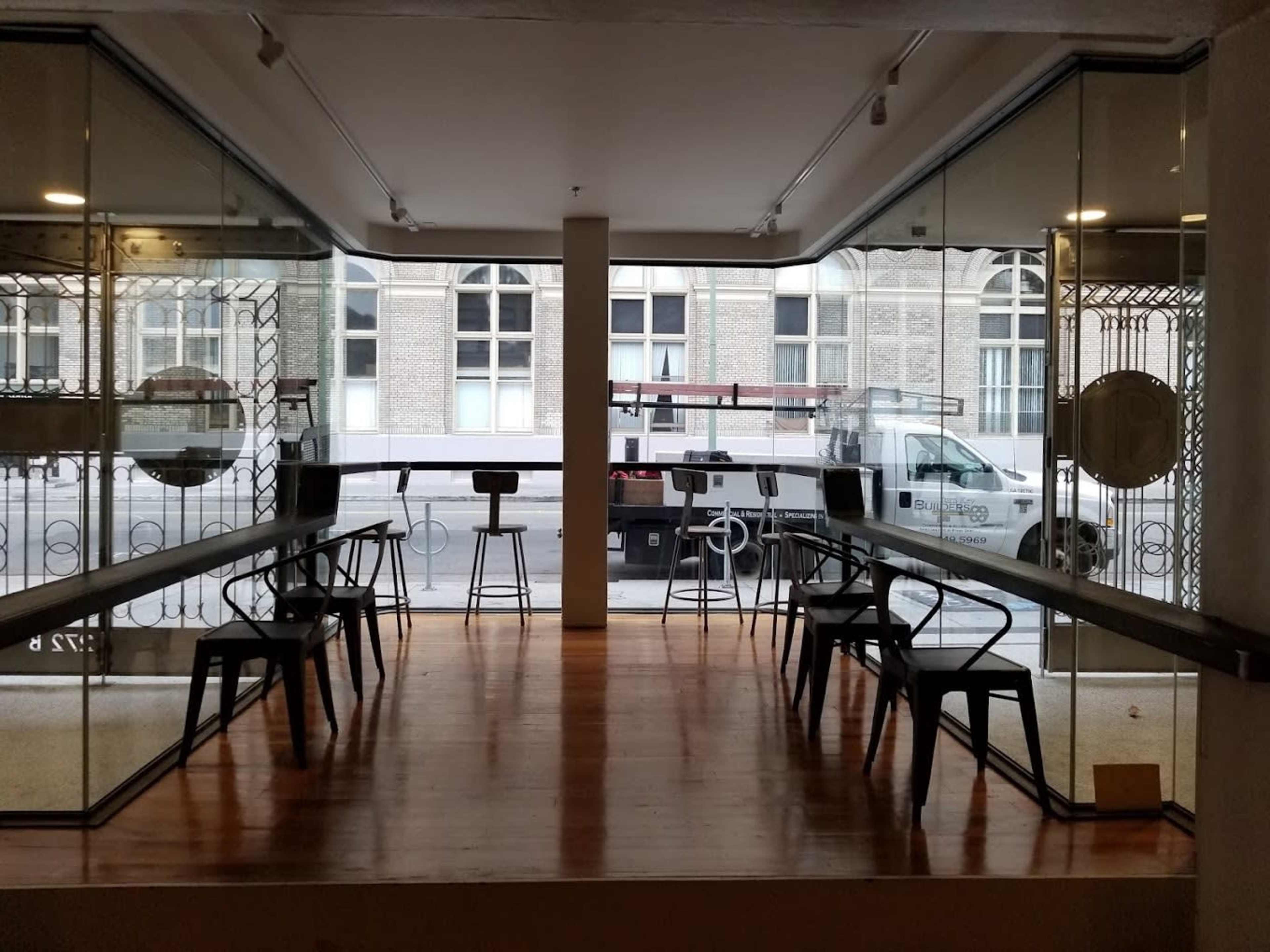The image shows a modern cafe interior featuring long wooden tables and metal chairs, with large glass windows providing a view of the street outside.