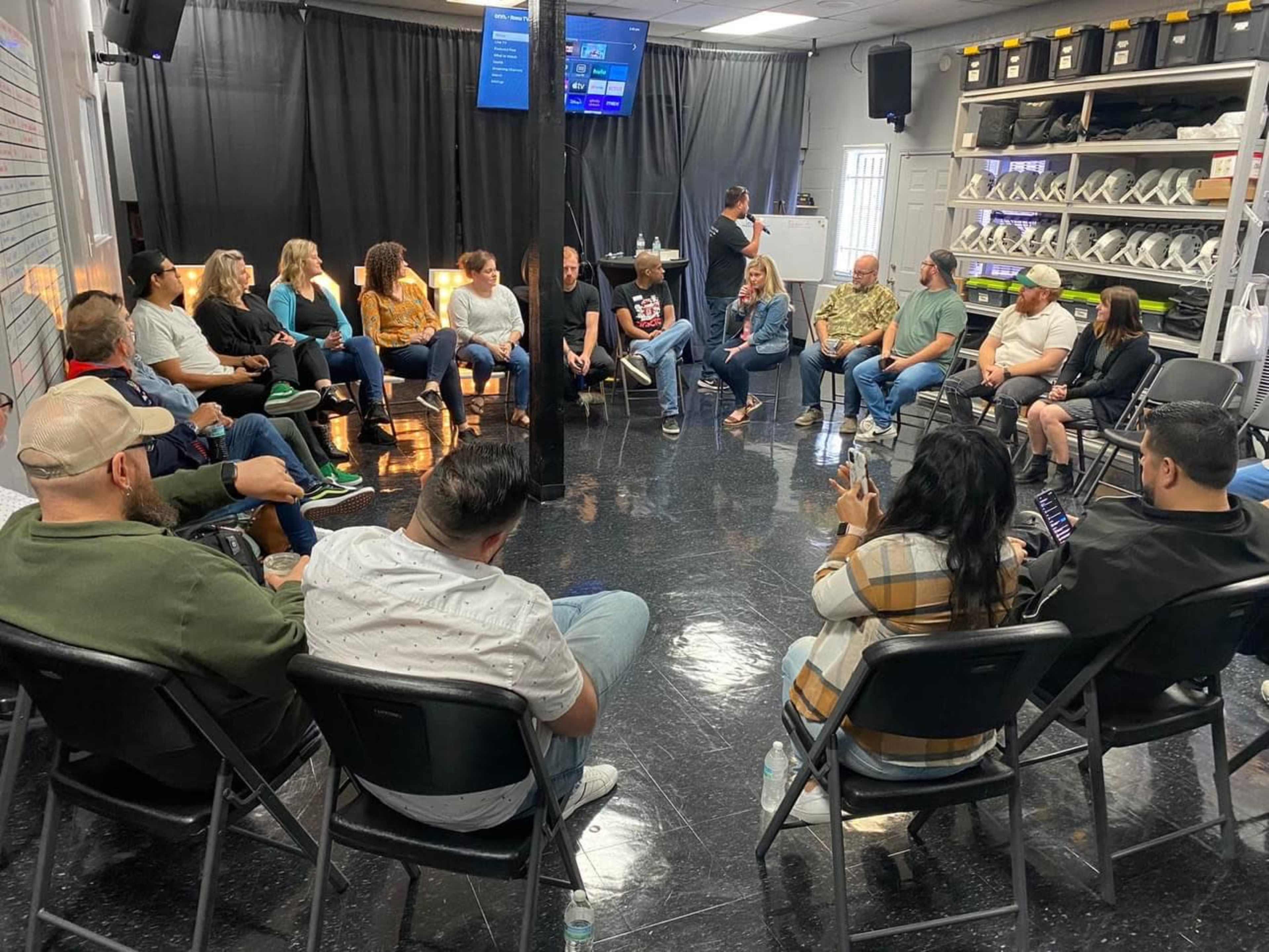 A group of people is seated in a circular arrangement in a well-lit meeting room with a presentation screen in the background.