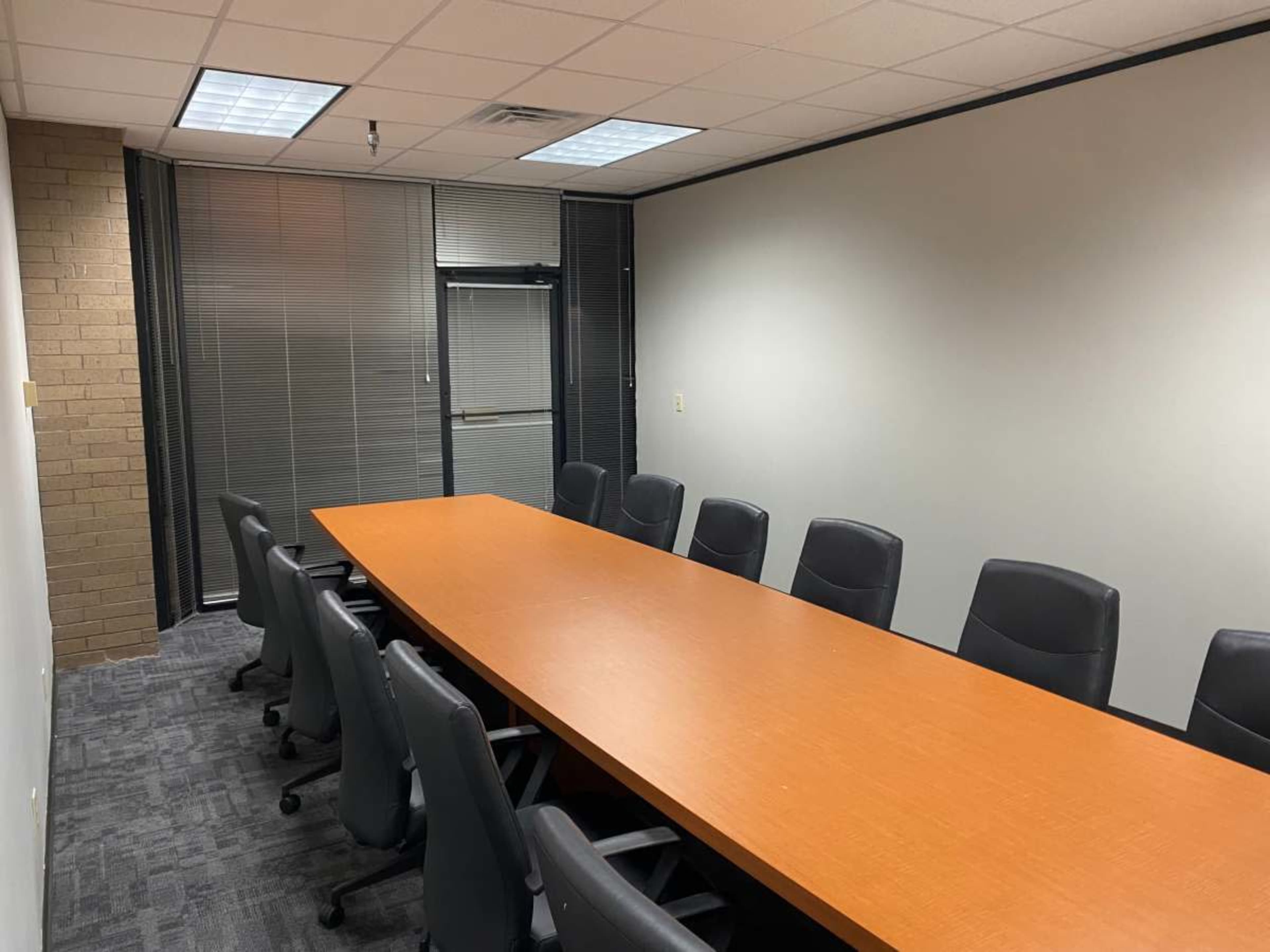 A conference room with a long wooden table surrounded by black chairs and windows covered with blinds.